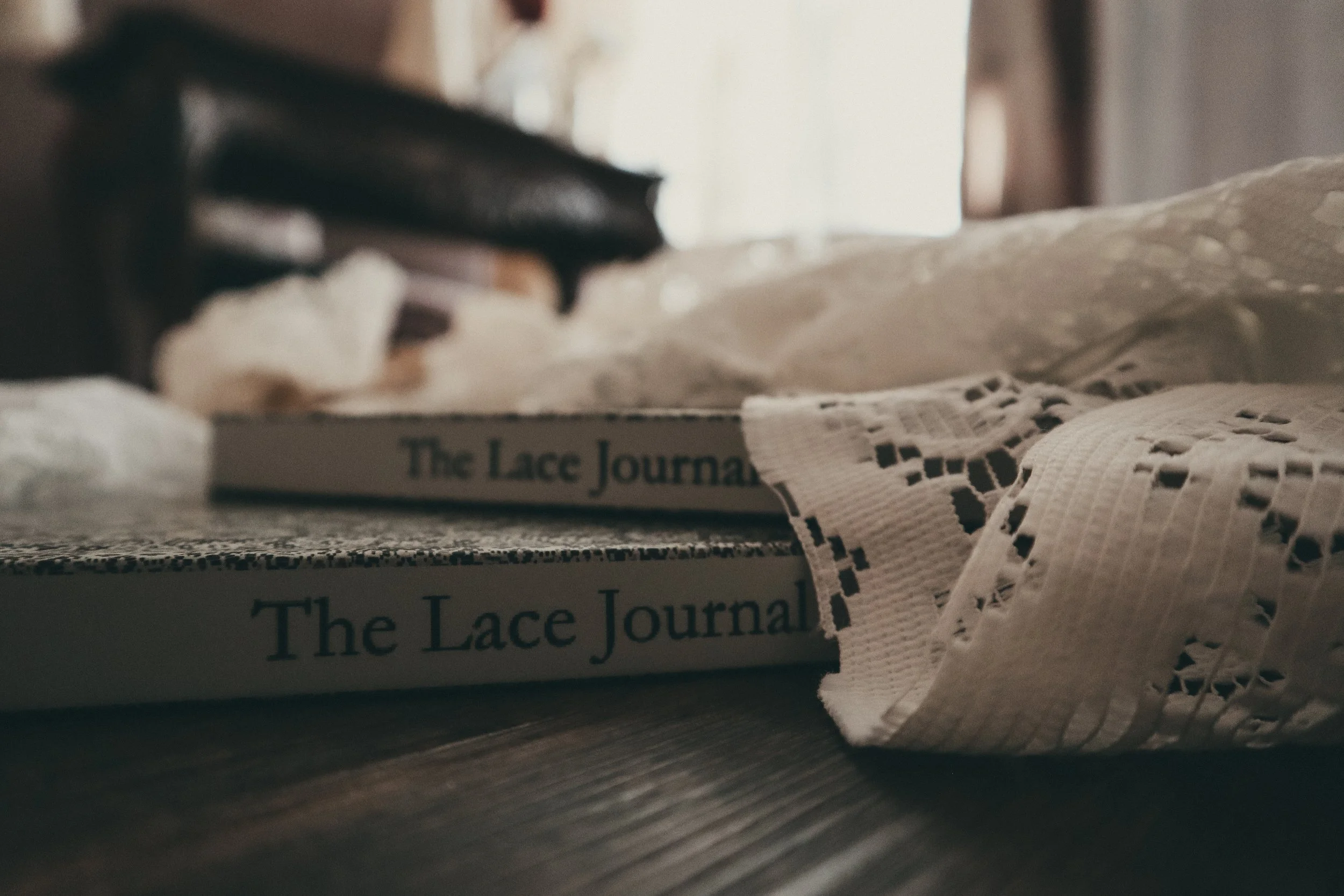 Close-up of a book titled 'The Lace Journal' with a white lace cloth partially covering it, placed on a wooden surface.