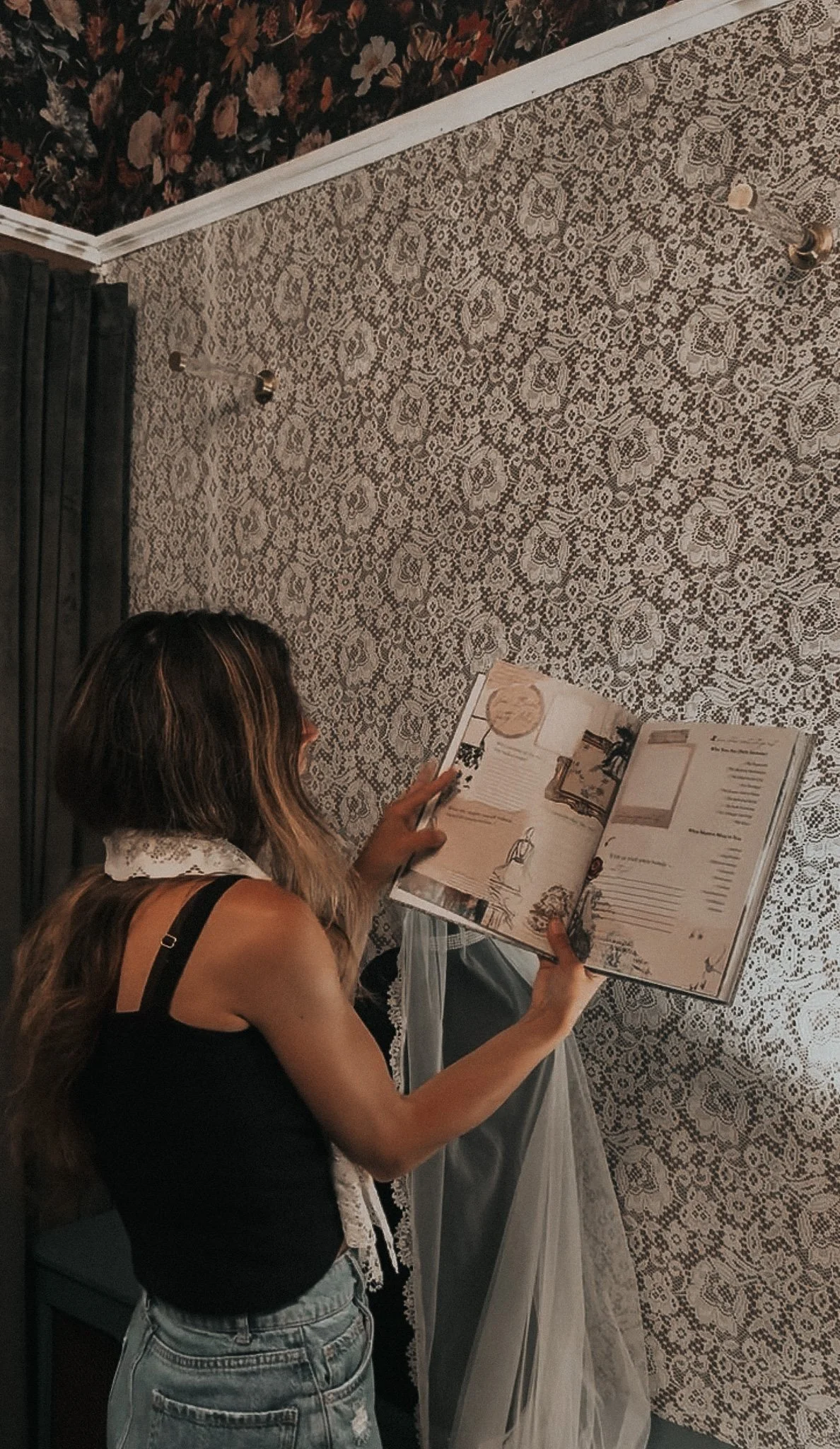 A woman with long, wavy brown hair reading a magazine while sitting on a gray chair in a room with floral-patterned wallpaper and a lace floral curtain.