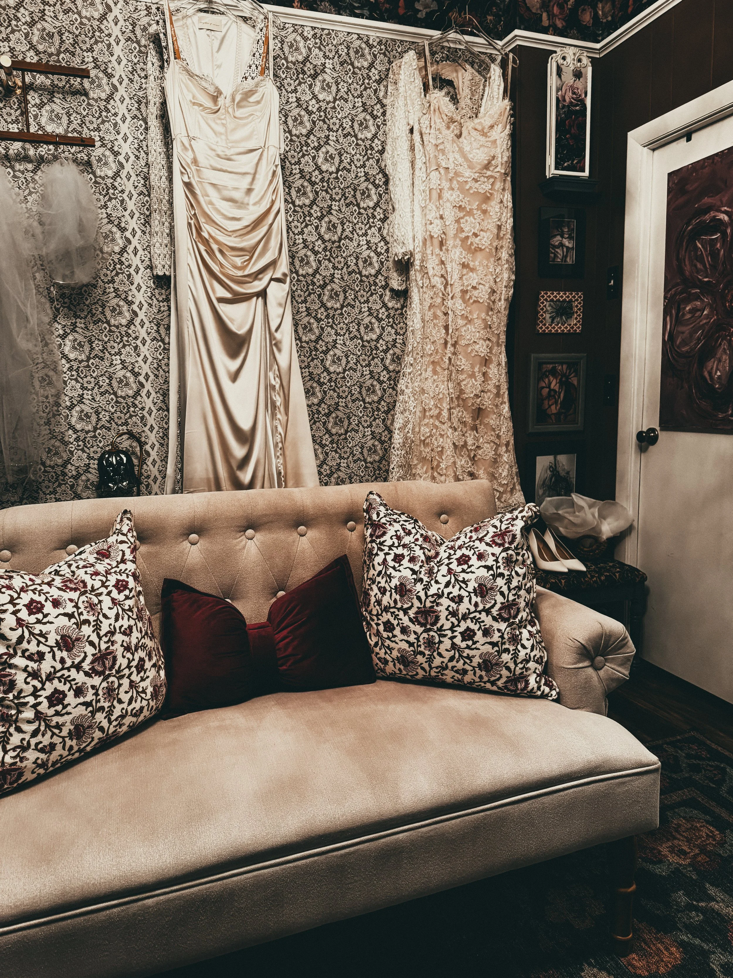 A beige sofa with four decorative pillows, two with floral patterns and two solid burgundy. Behind the sofa, two wedding dresses hang on a lace-covered wall. A pair of white shoes sits on a small table beside the sofa, and the room features dark walls with framed artwork.