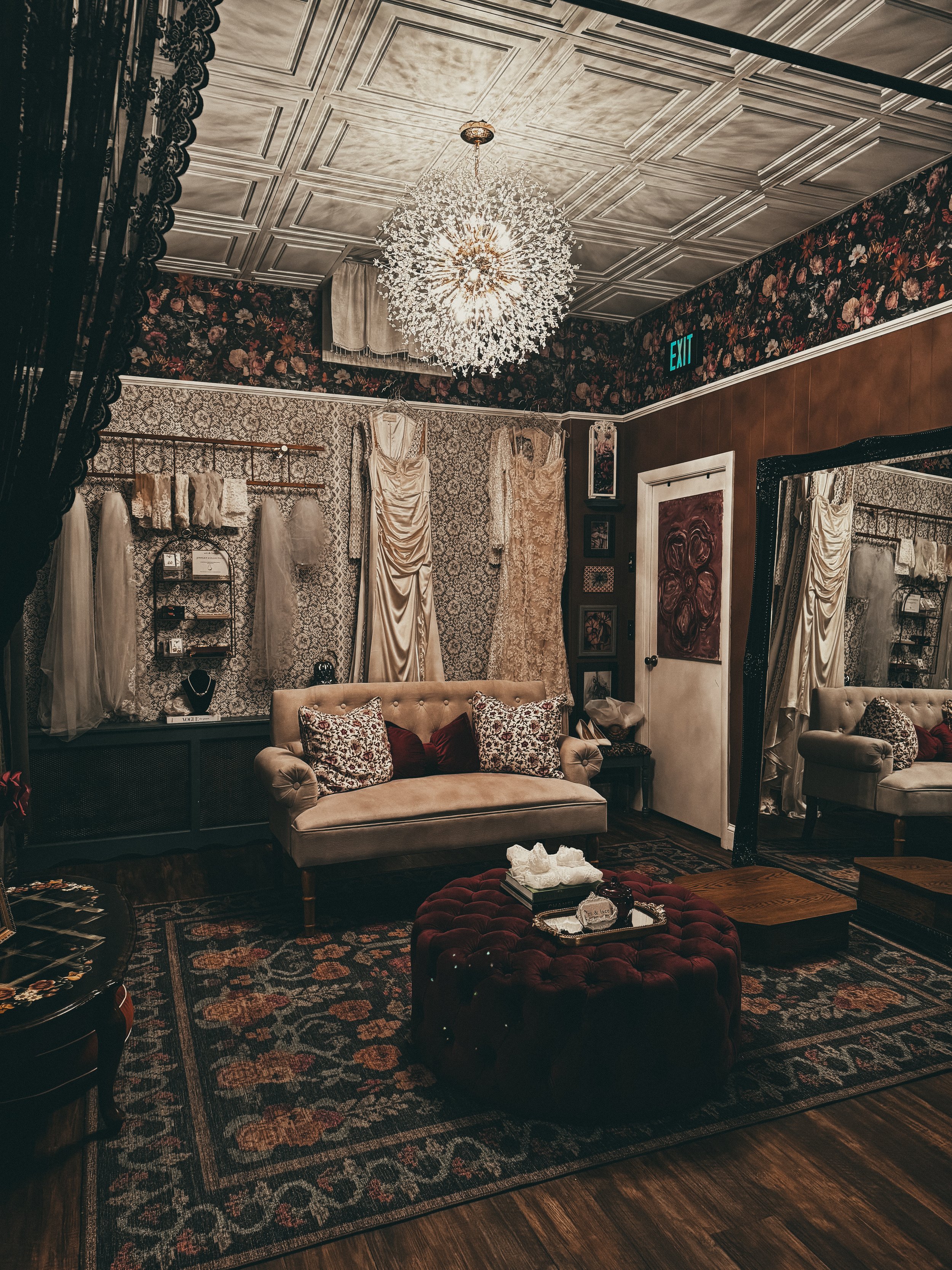 A cozy vintage-inspired boutique living room with floral wallpaper, cream-colored sofa, large mirror, and decorated with wedding dresses and accessories, chandelier lighting, and a patterned rug.