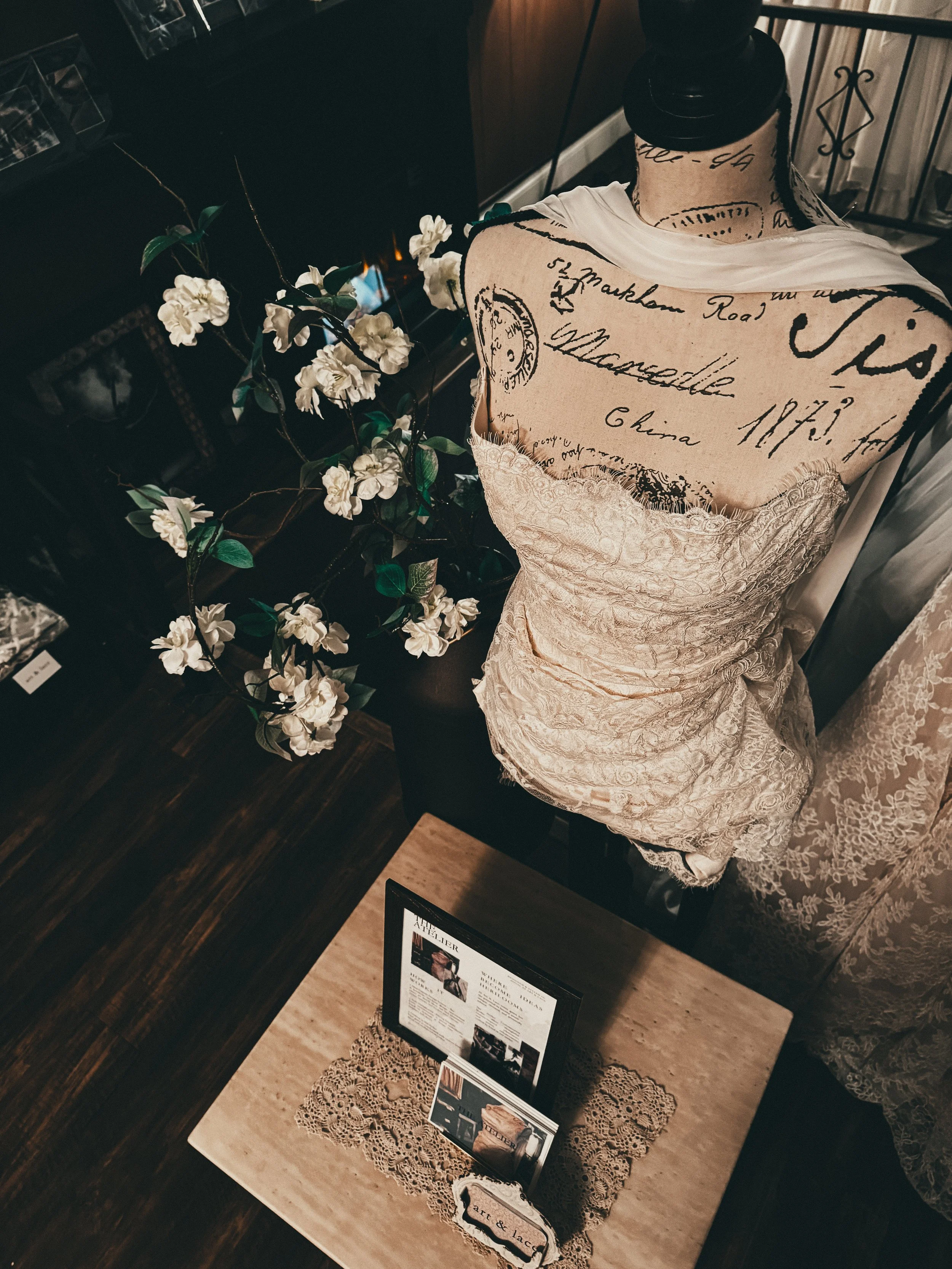 Display of a mannequin dressed in vintage-style lace dress with handwritten script fabric covering the shoulders, next to a branch of white artificial flowers on a dark wood surface, informational framed art & lace doily on a light-colored table.