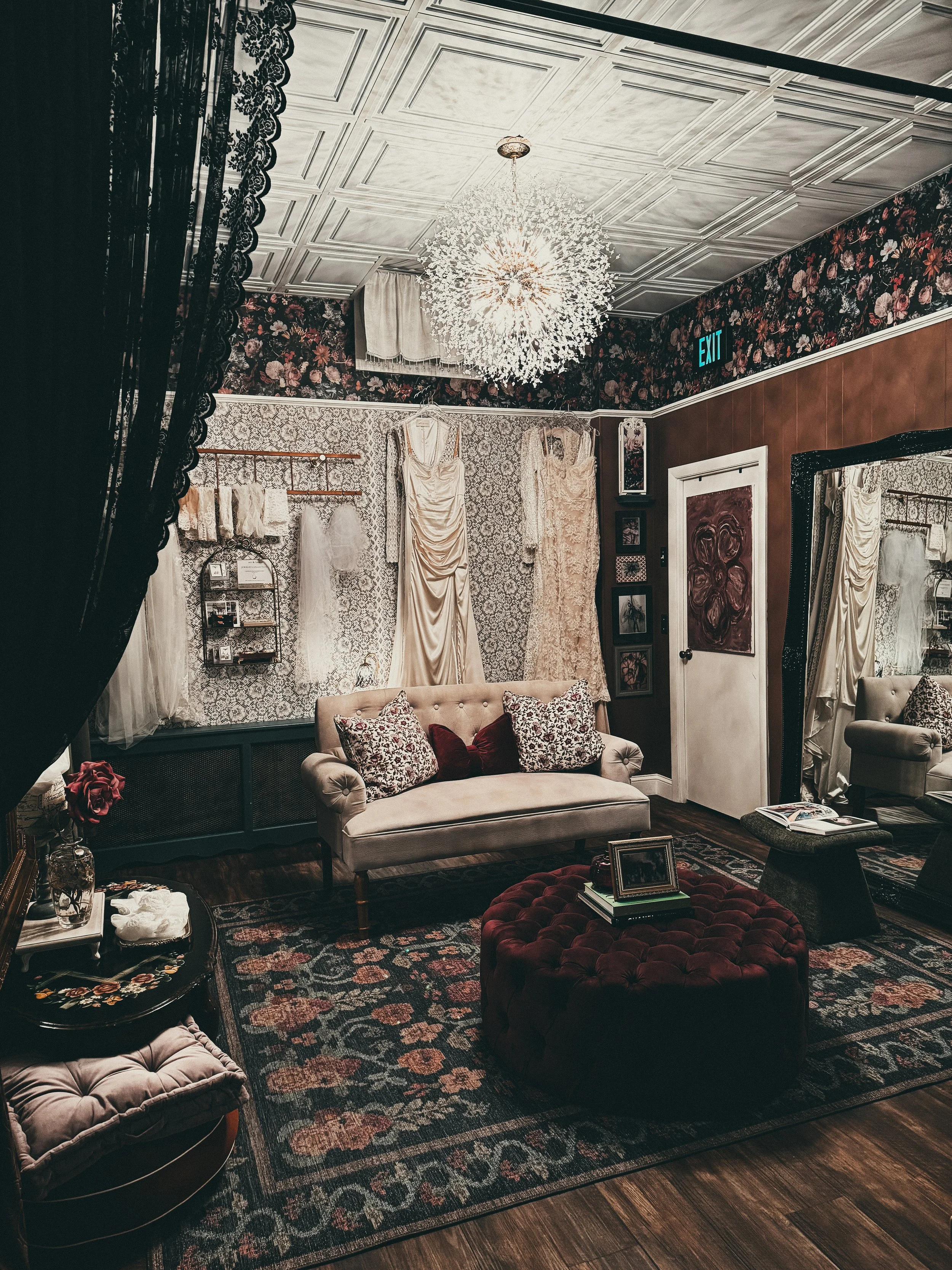 Vintage-inspired dressing room with floral wallpaper, lace curtains, and a tufted beige sofa with decorative pillows, a tufted round ottoman, framed artwork, and a large ornate mirror.