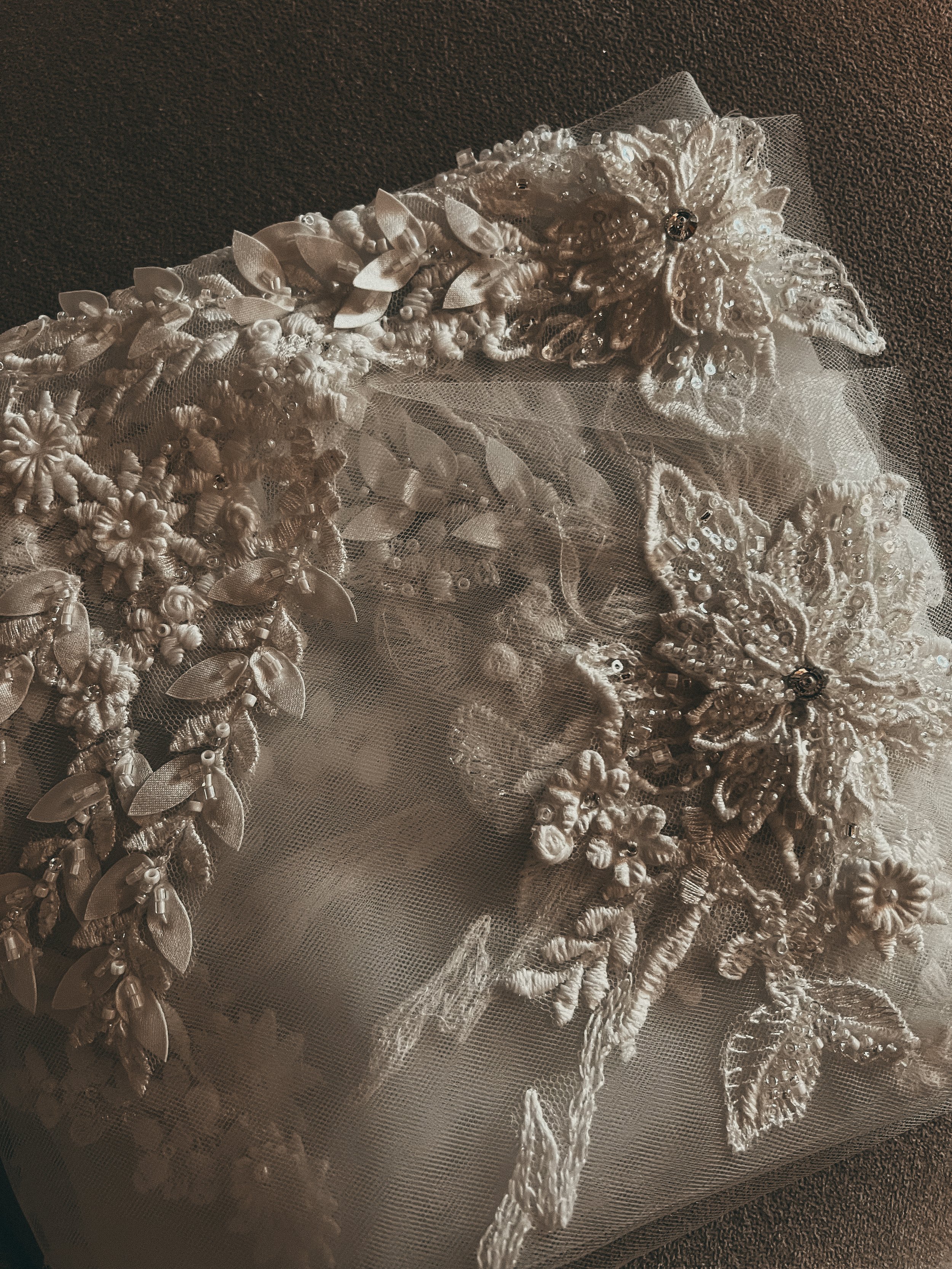 A white, intricately embroidered bridal veil with floral and butterfly designs and bead embellishments on a dark surface.