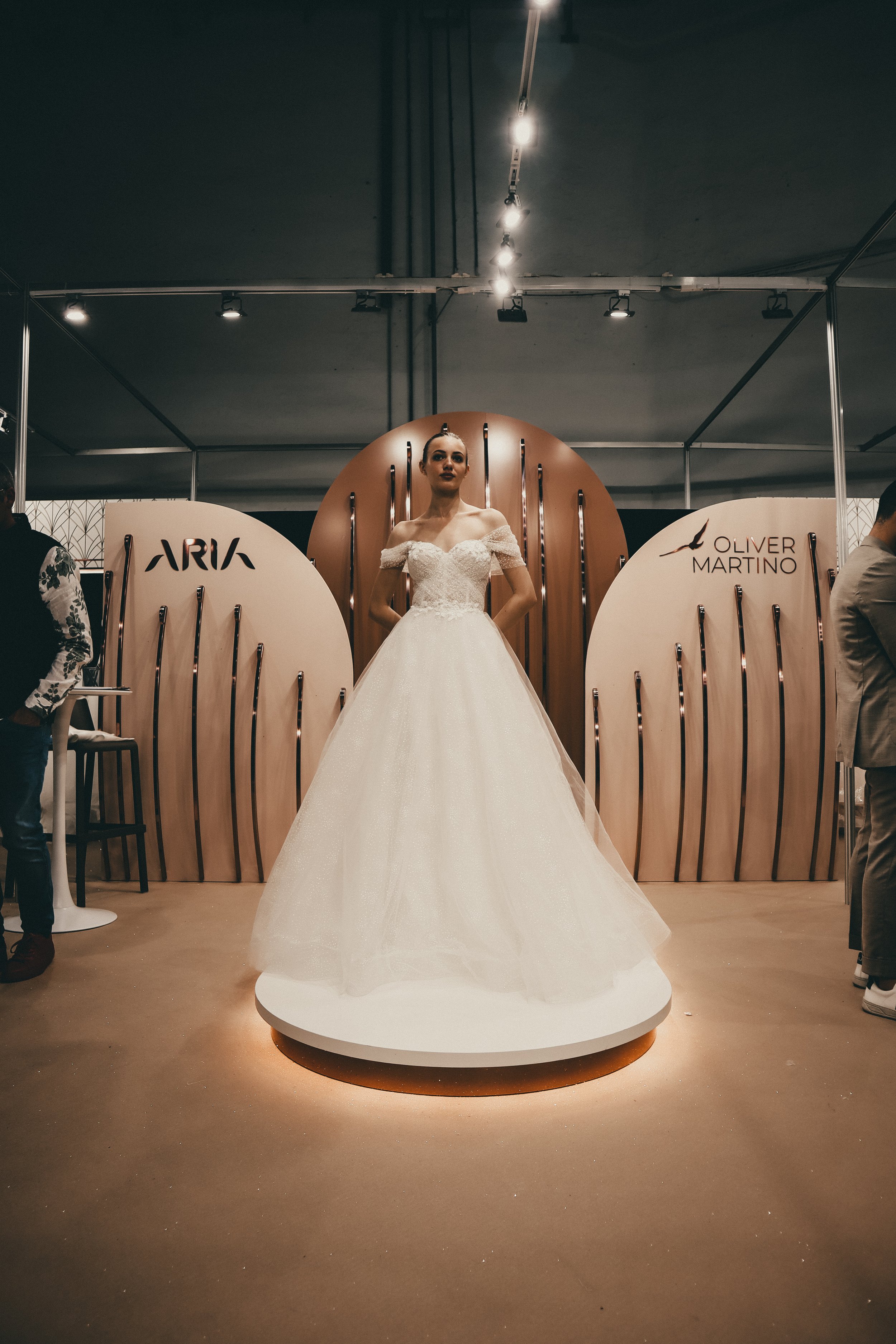A mannequin in a wedding dress displayed on a circular platform at a wedding expo, with decorative panels behind it featuring the brands 'ARIKA' and 'OLIVER MARTINO'.