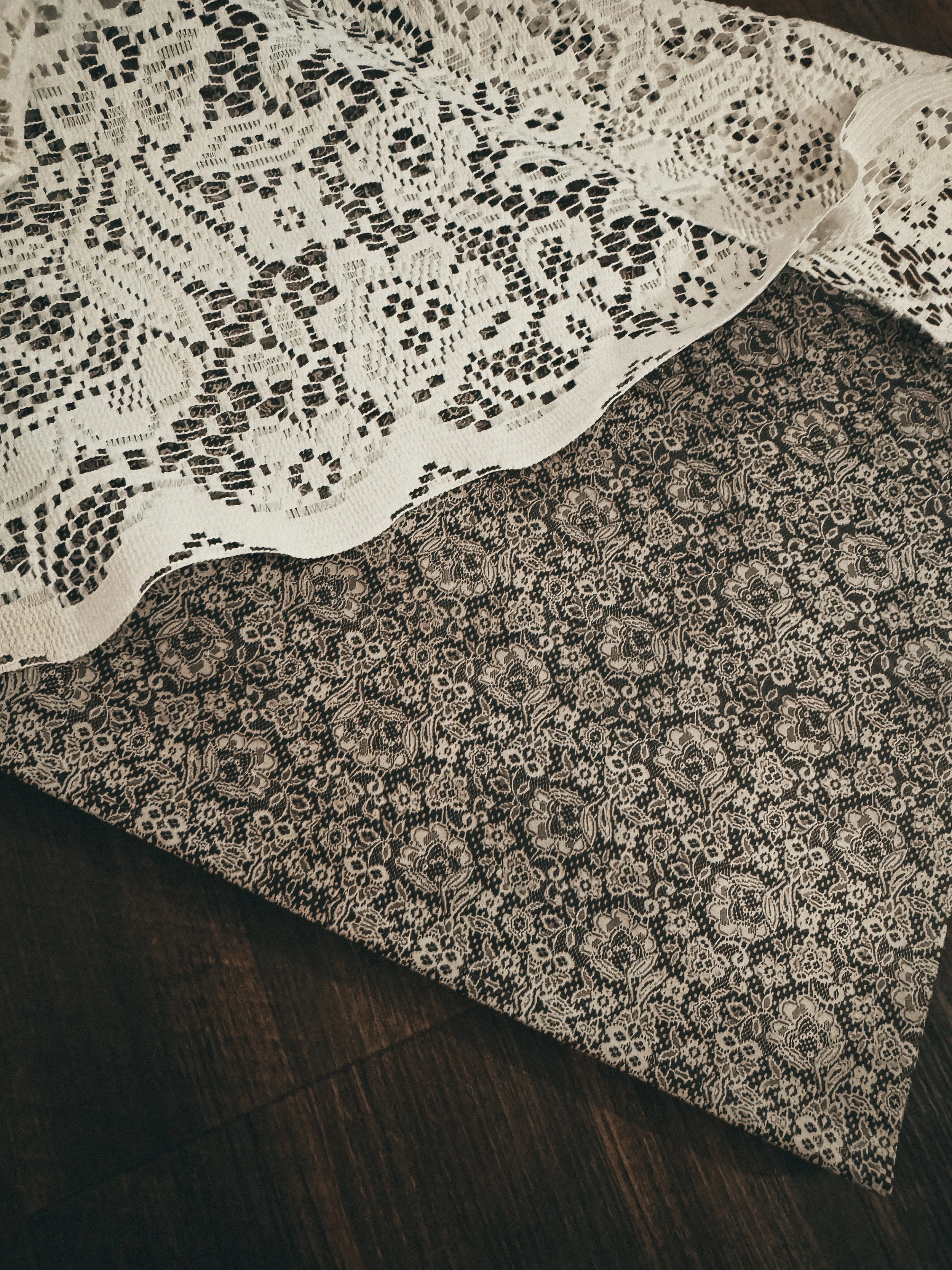 A close-up of a piece of fabric with an intricate black lace pattern on a light background, resting on a dark wooden surface.