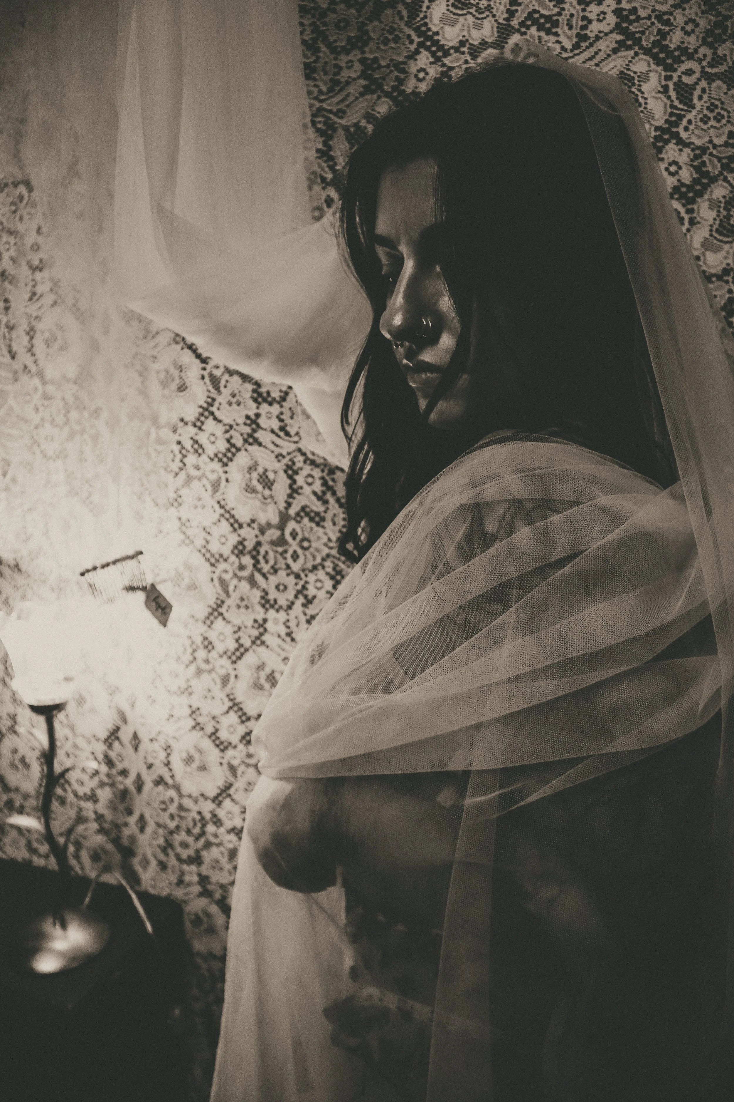 A woman with dark hair and a nose piercing in a contemplative pose, wearing a sheer veil, standing against a lace-patterned wall with a small table and lamp nearby.