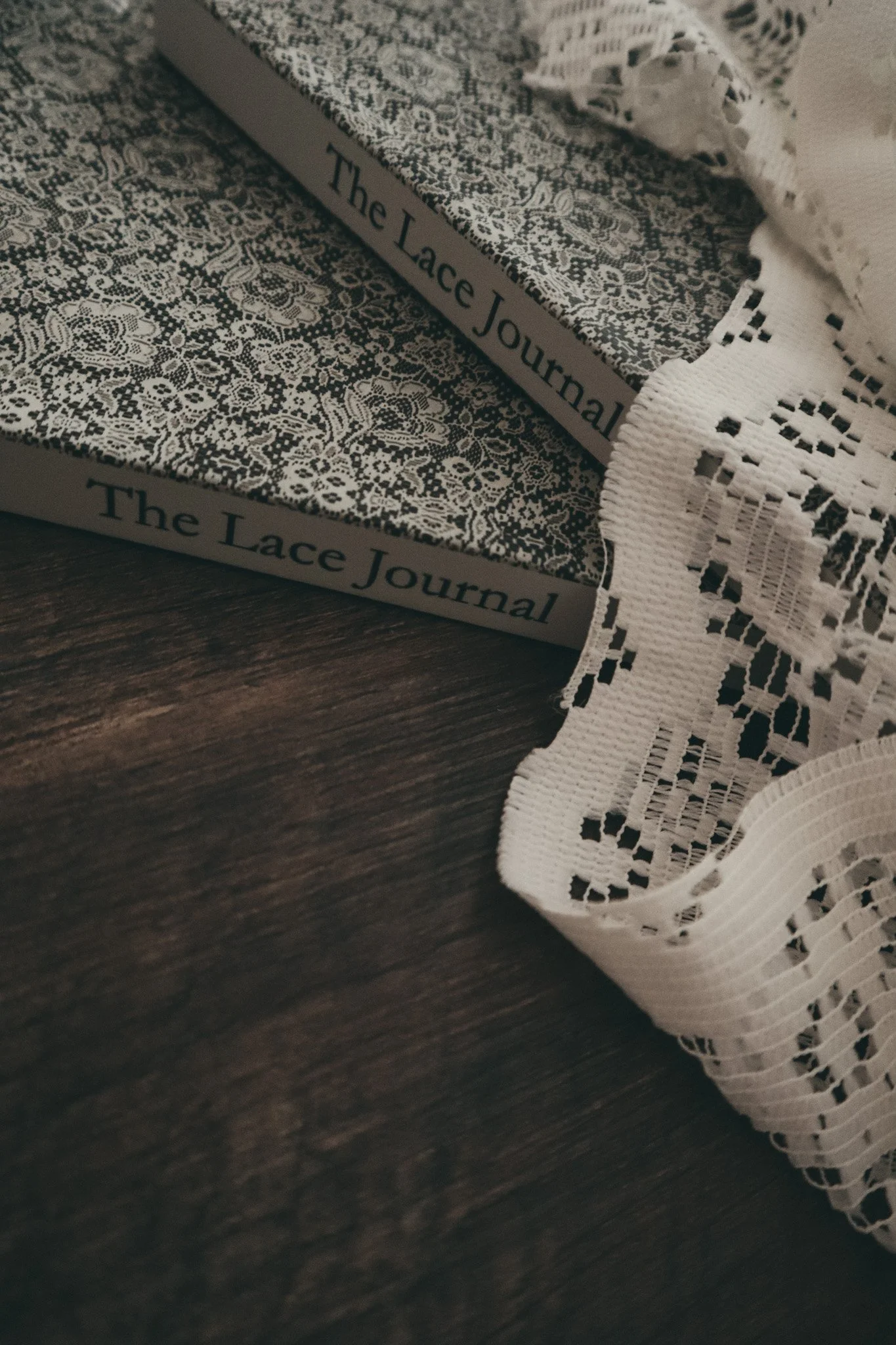Close-up of a book titled 'The Lace Journal' with intricate lace-patterned pages and a white lace fabric draped over it, on a dark wooden surface.