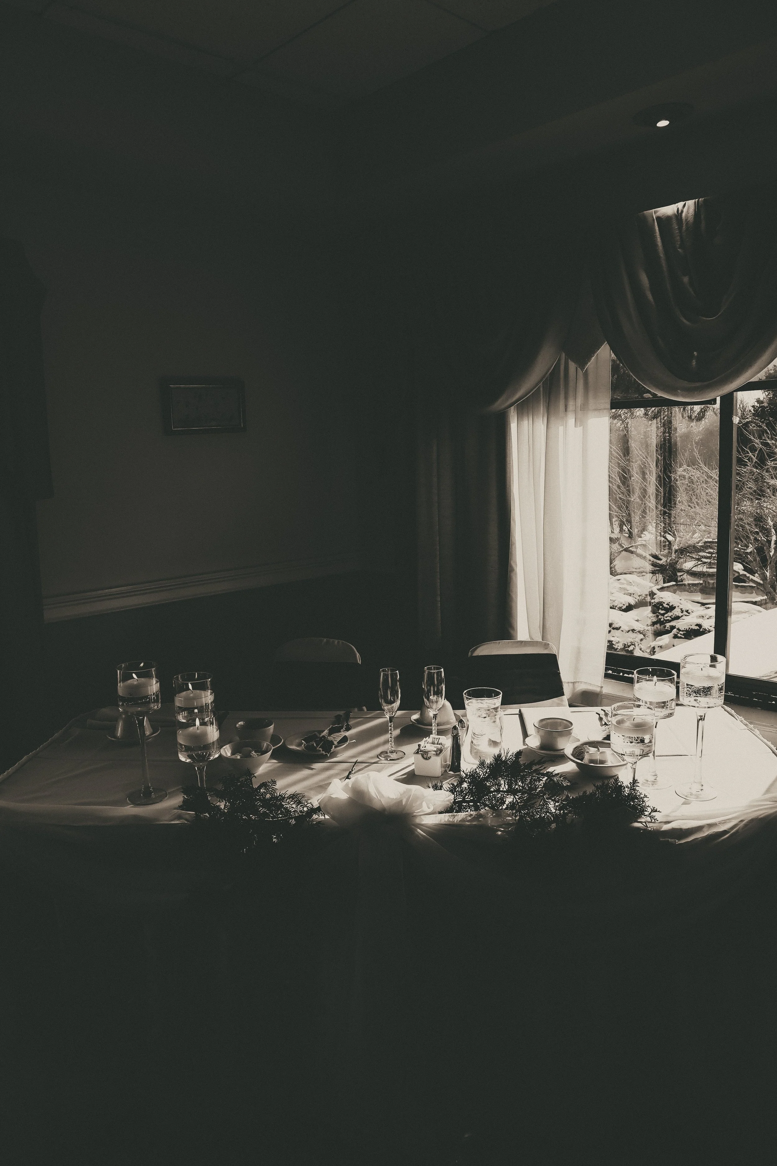 A dimly lit dining room with a table set with glasses, bowls, and decorative greenery, with a large window revealing a snowy outdoor landscape.