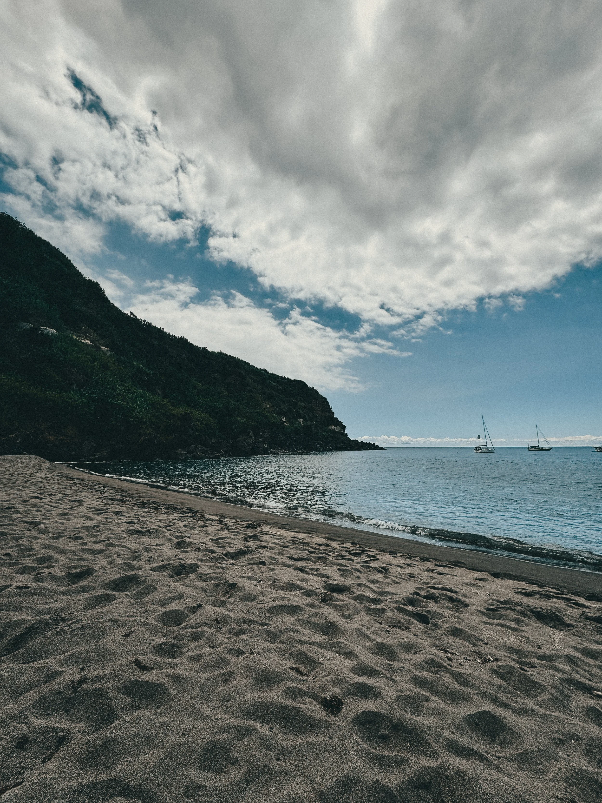 A sandy beach with footprints, calm ocean waters, sailboats, a mountain on the left, and a partly cloudy sky.