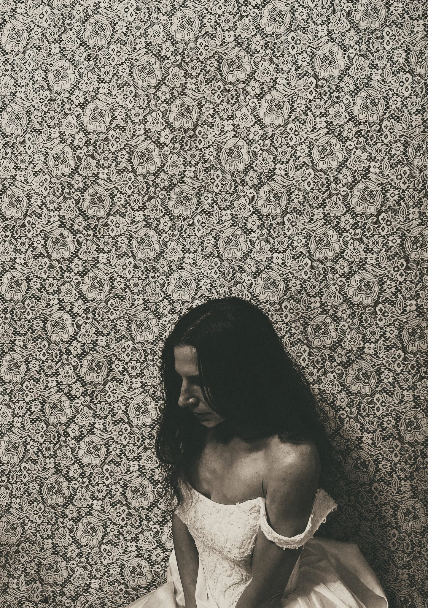A moody black-and-white portrait of a woman in an off-the-shoulder vintage wedding gown, seated against a lace wall. Her head is lowered, creating a somber, introspective atmosphere.