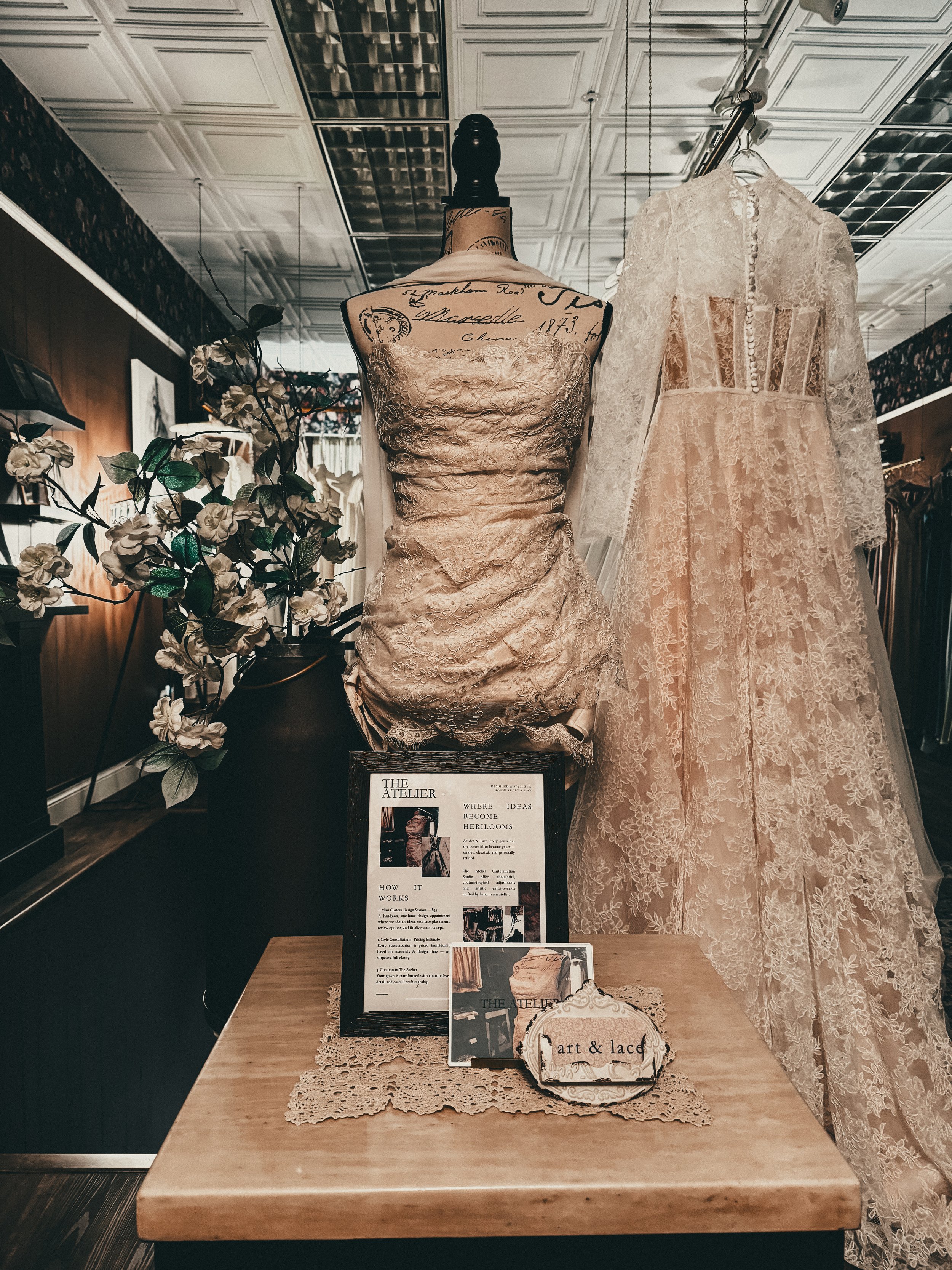 Display of vintage lace dress on mannequin and lace wedding gown on hanger in a boutique with informational sign on table.