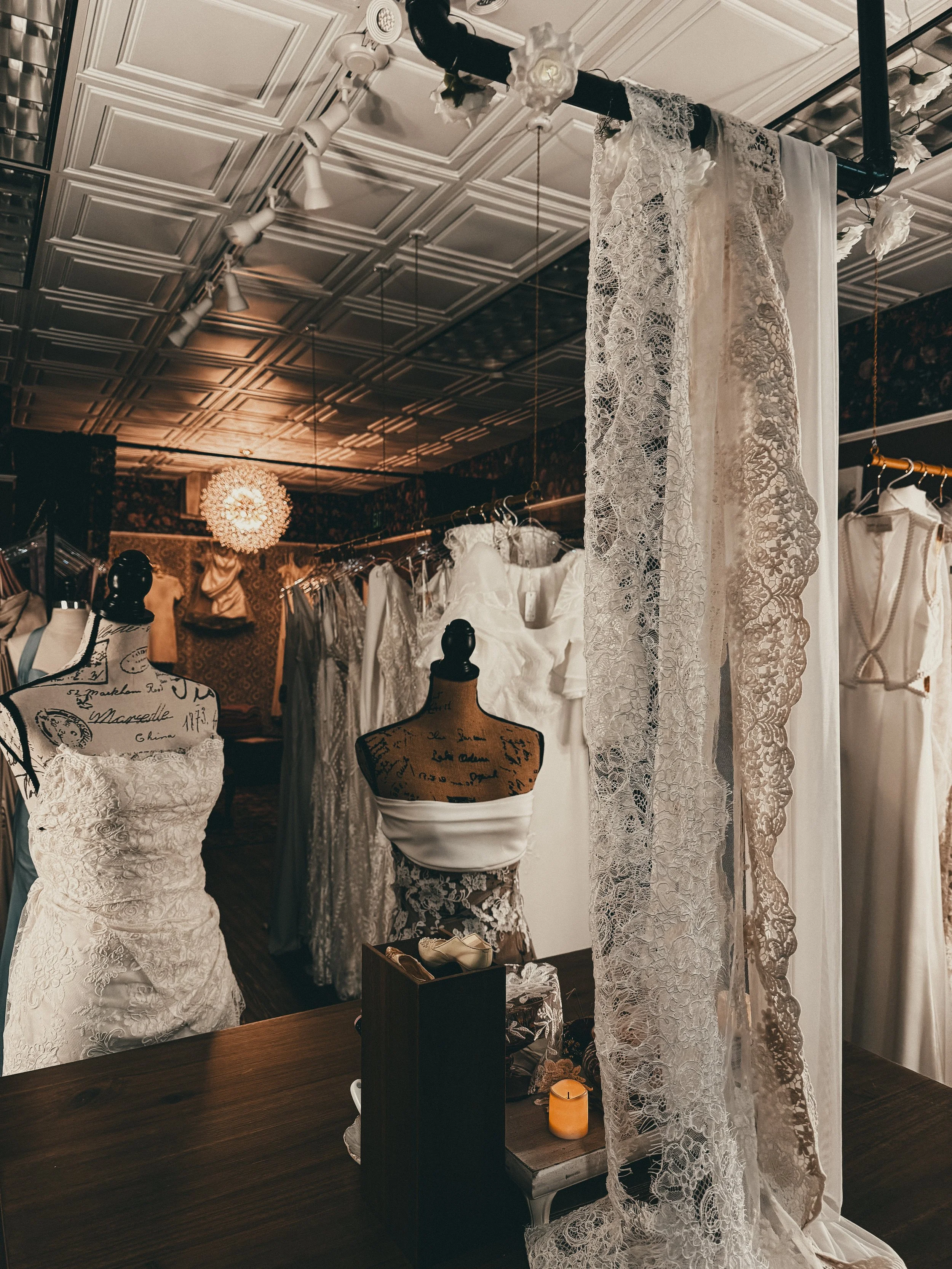 Display of wedding dresses in a boutique, with lace, satin, and gown styles, on mannequins and hangers, illuminated by warm lighting.