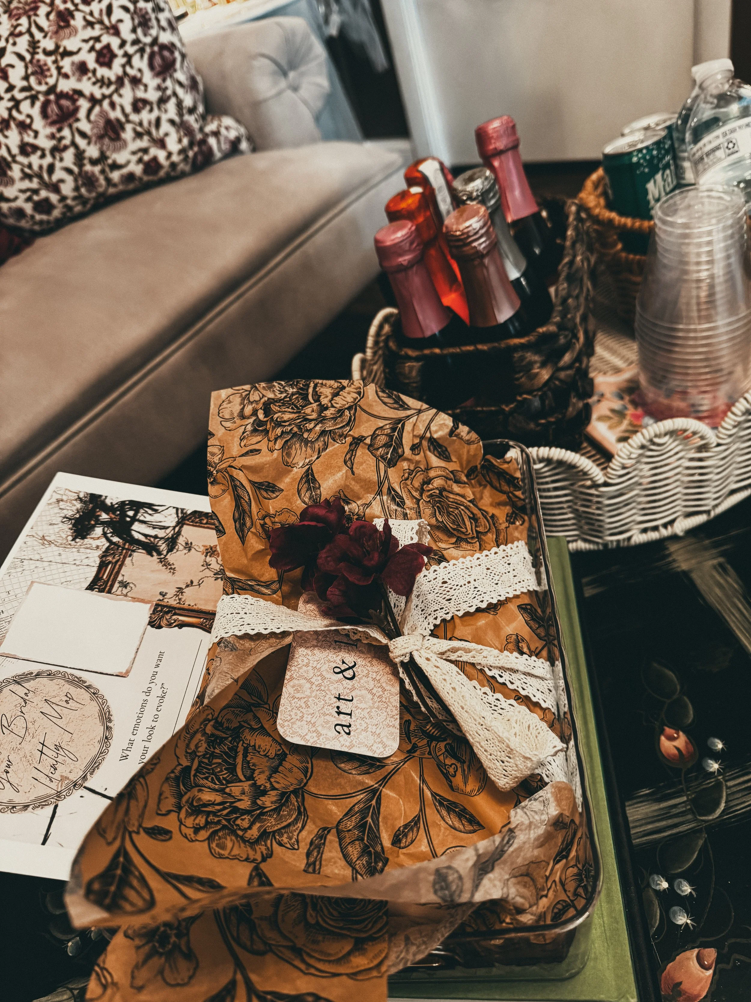 Basket of bottled beverages and clear plastic cups on a table, with a patterned paper bag, greeting card, and snacks nearby.