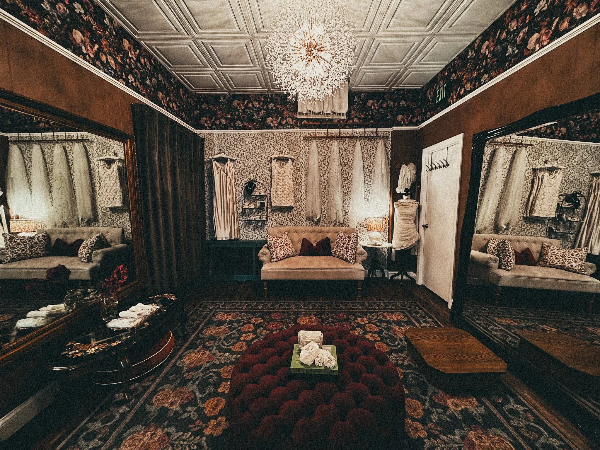 A vintage dressing room with floral wallpaper, mirrors, and a seating area with an ottoman. The room features ornate ceiling tiles, a chandelier, and various clothing and accessories displayed along the walls.