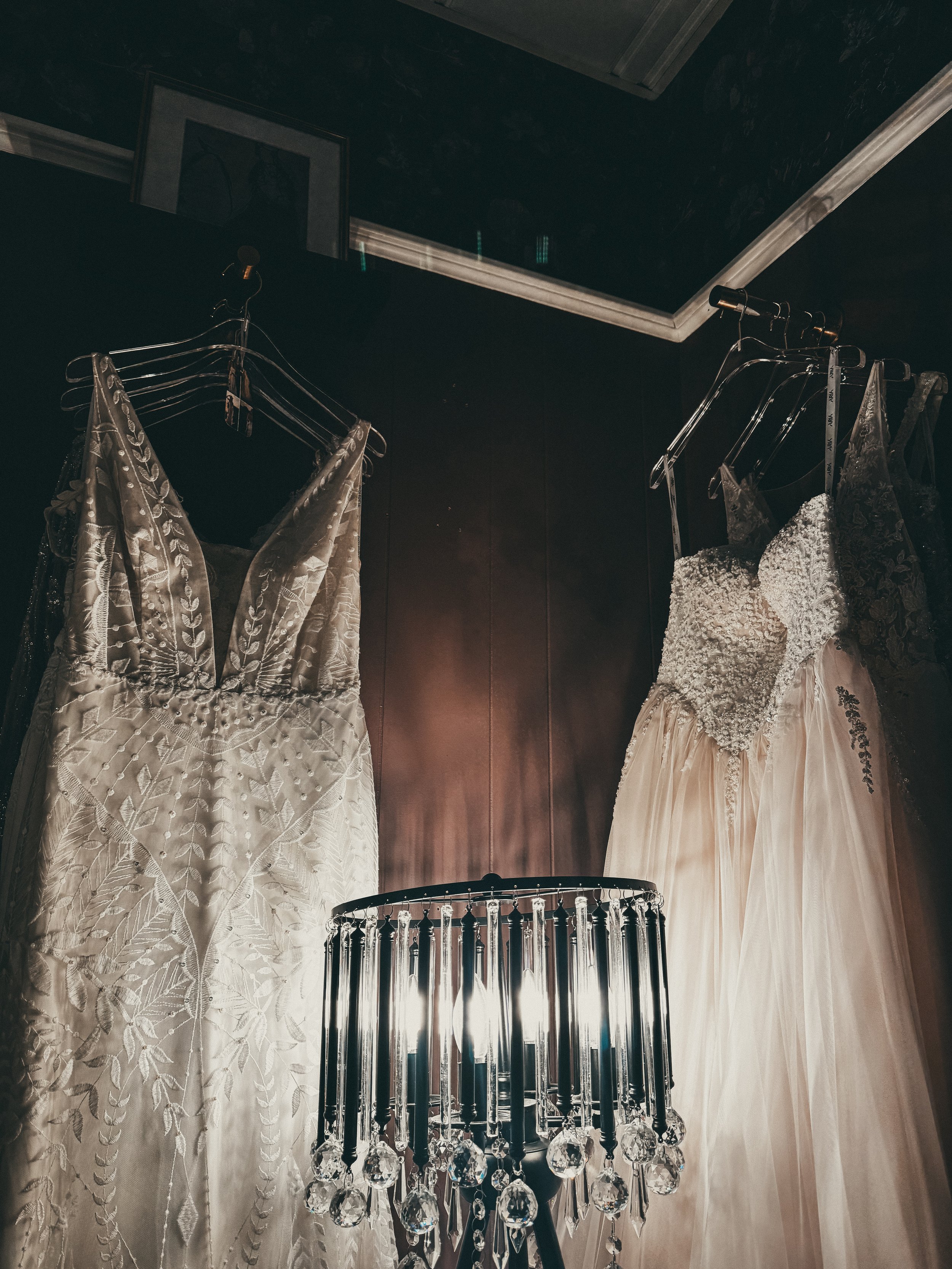 Two wedding dresses hanging on a dark wall with a small chandelier in the foreground.