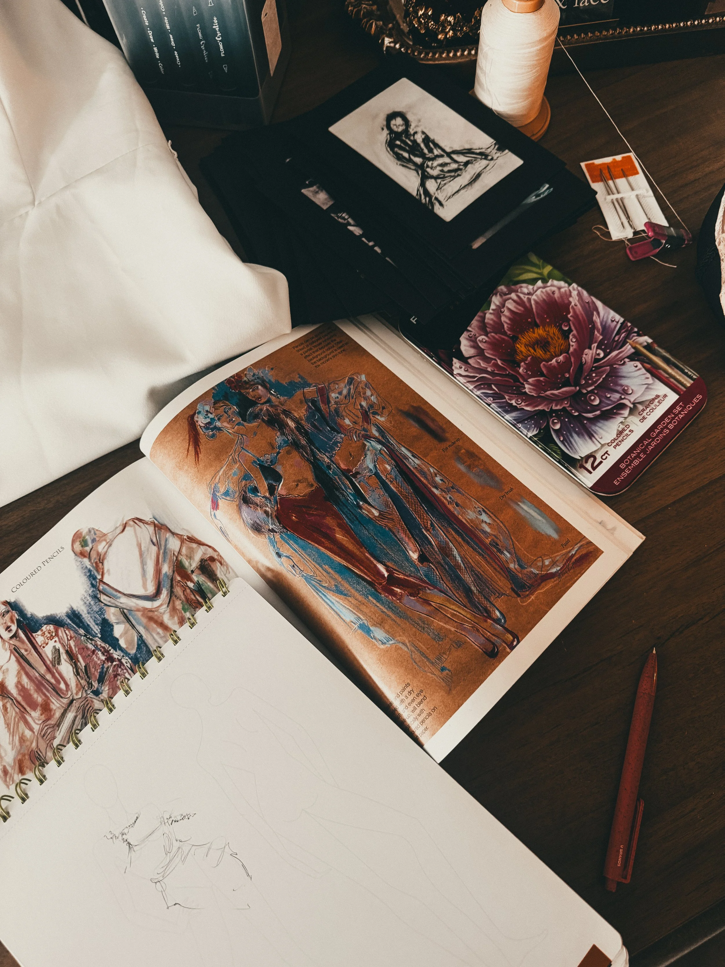 A table with art supplies including a sketchbook with pencil drawings, an open magazine with fashion illustrations, a color palette with a flower photo, a pen, a spool of thread, and sewing accessories.