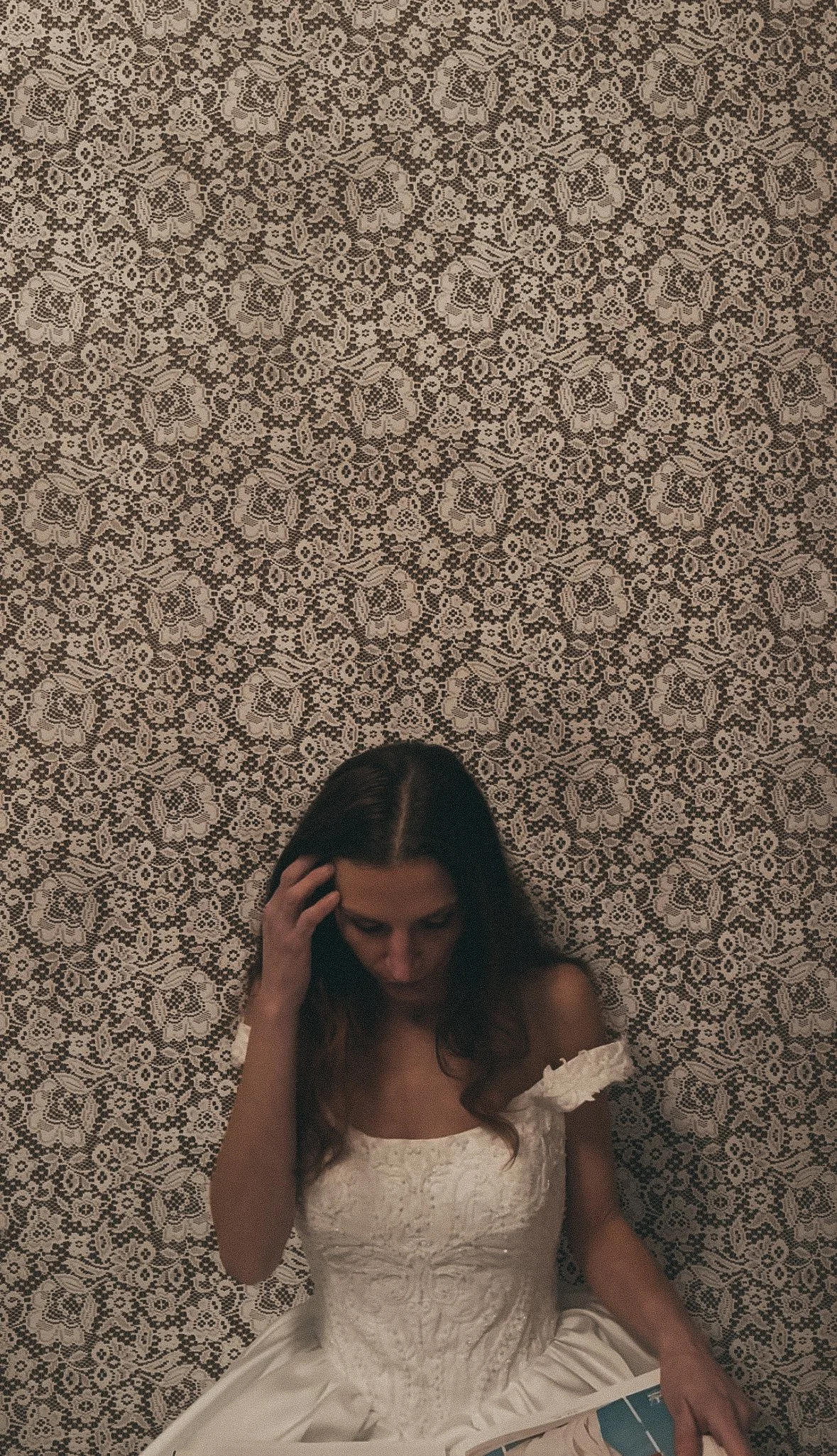 A woman in a white dress sits on a patterned carpet, looking down with her hand on her head.