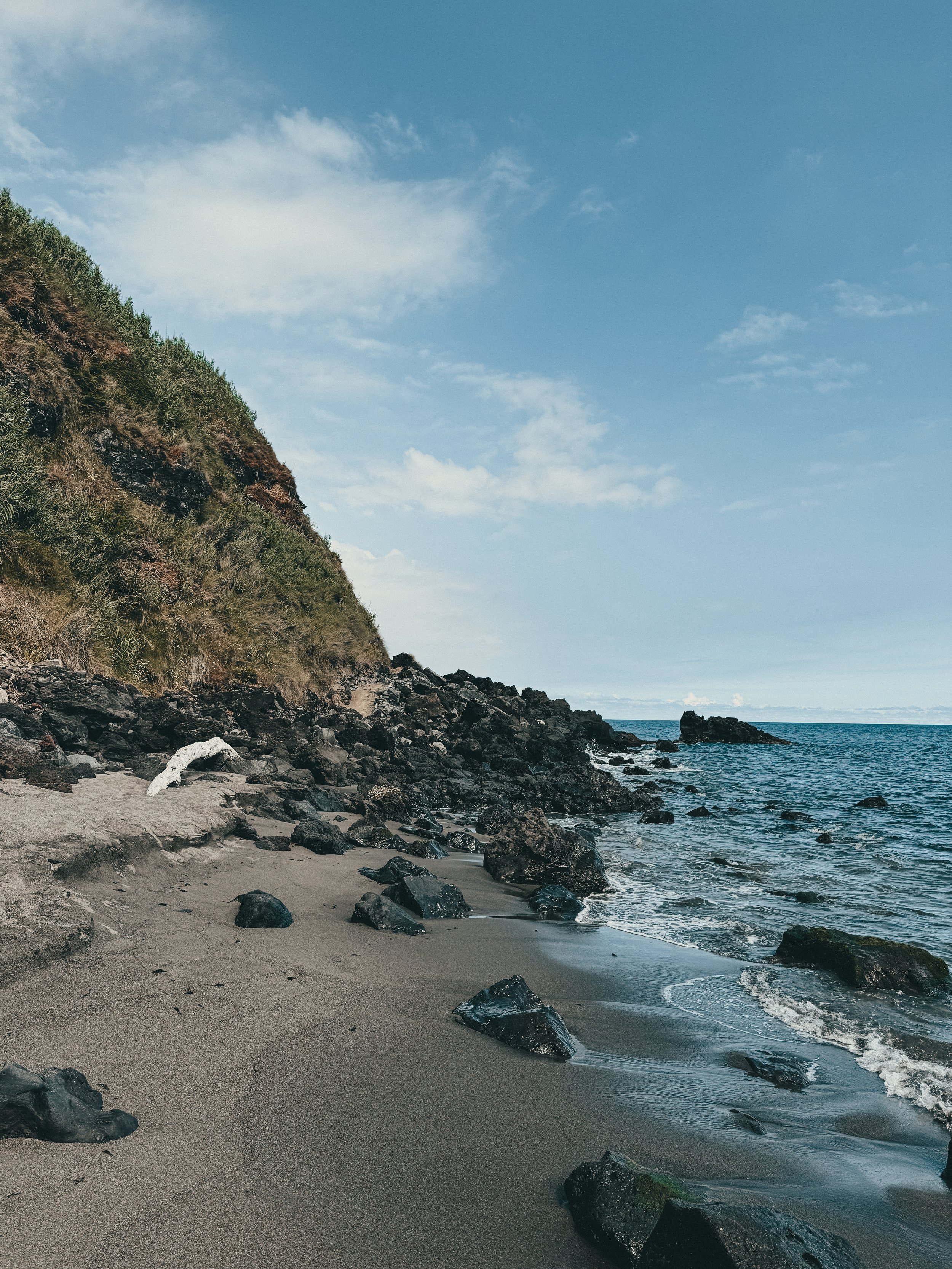 A rocky beach with dark sand, a hillside covered in greenery on the left, and an ocean with small waves on the right under a blue sky with scattered clouds.