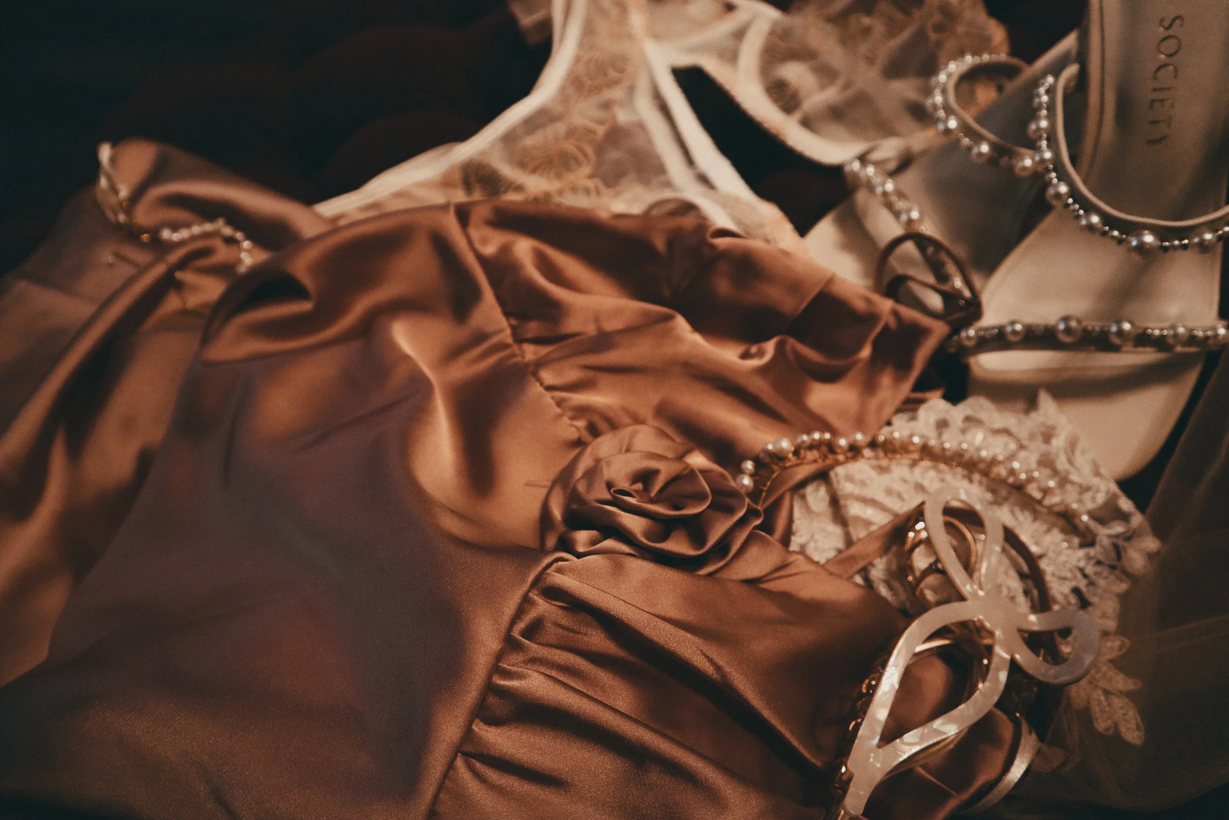 Close-up of a dressing table with accessories, including a satin brown garment, pearl necklaces, and sunglasses, all in warm ambient lighting.