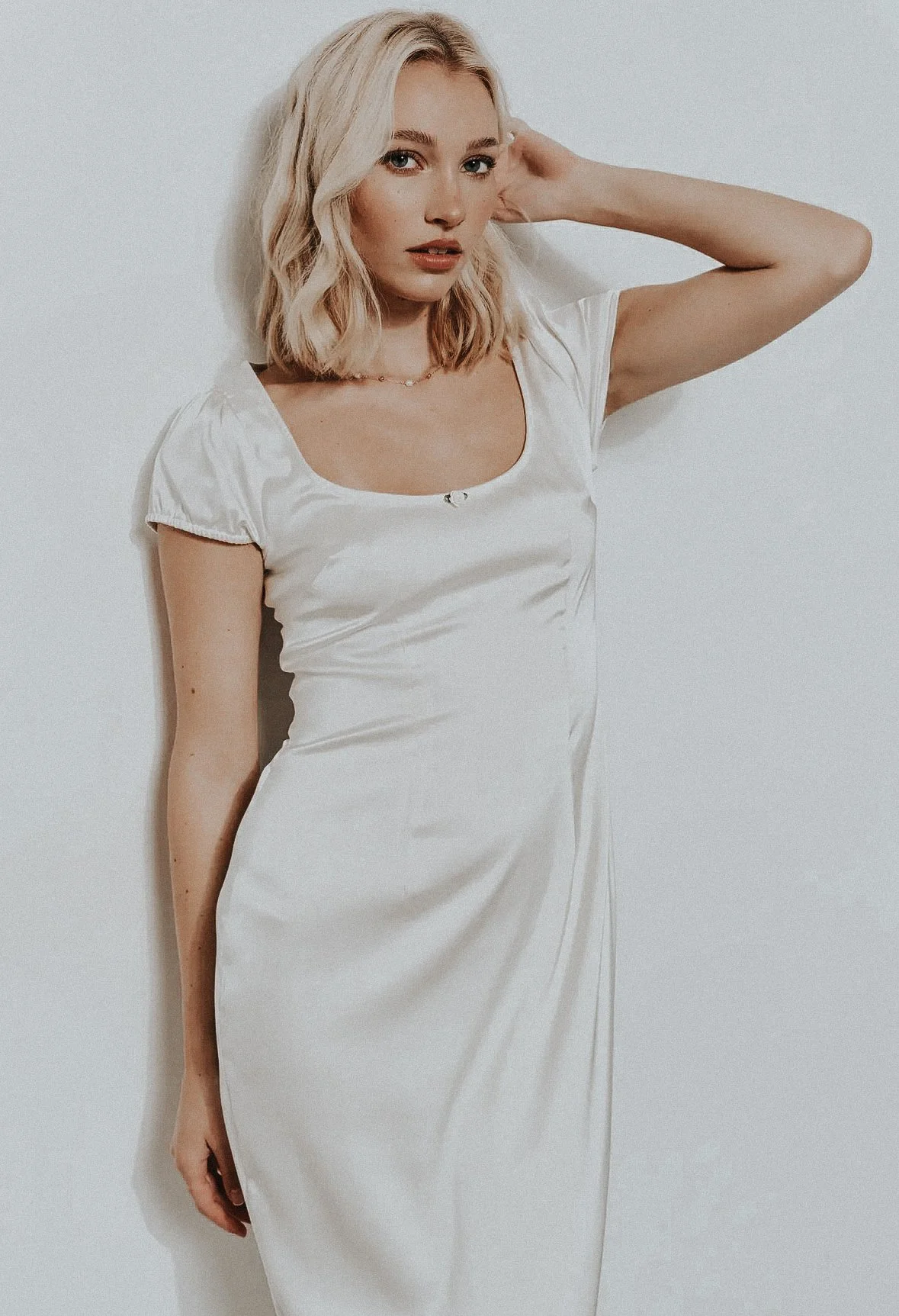 A woman with blonde hair and fair skin wearing a white dress, standing against a white wall.