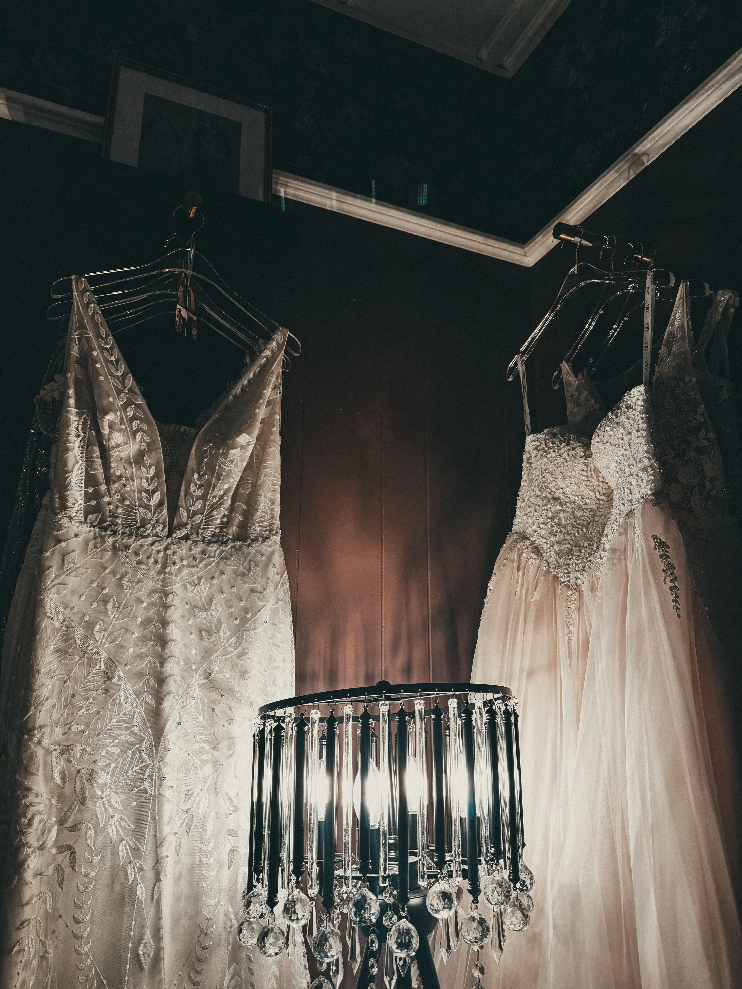 Two wedding dresses hanging on hooks inside a fitting room with dark walls and a decorative chandelier in the foreground.