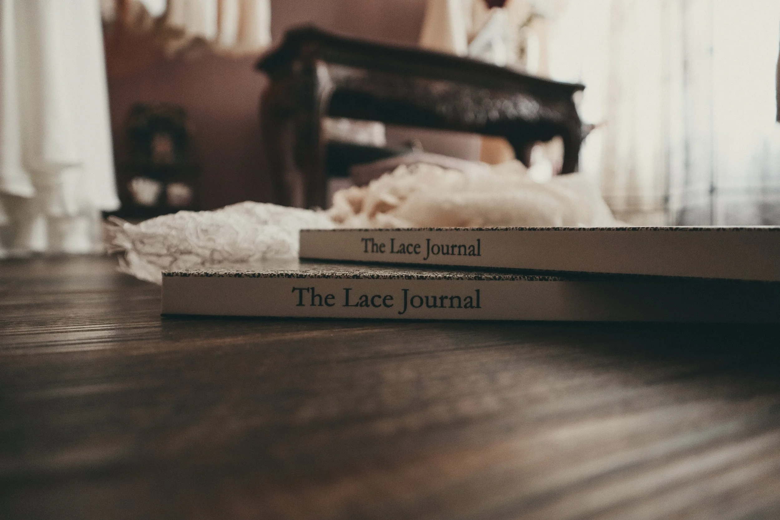 Two copies of a book titled 'The Lace Journal' placed on a wooden surface with lace fabric in front of them. In the background, there is a dark wooden table or cabinet and curtains to the right.