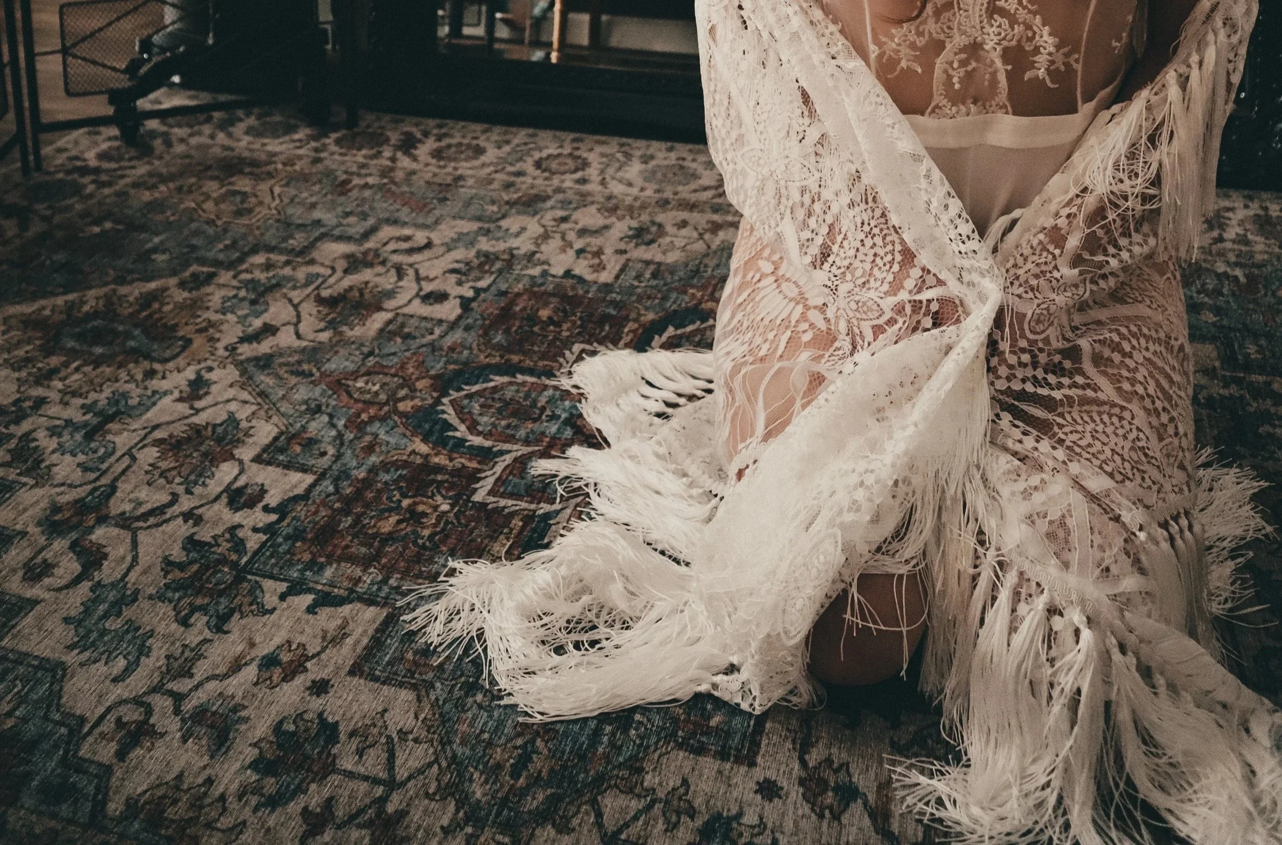 Close-up photo of a person sitting on a patterned rug, wearing a white lace dress with fringe details.