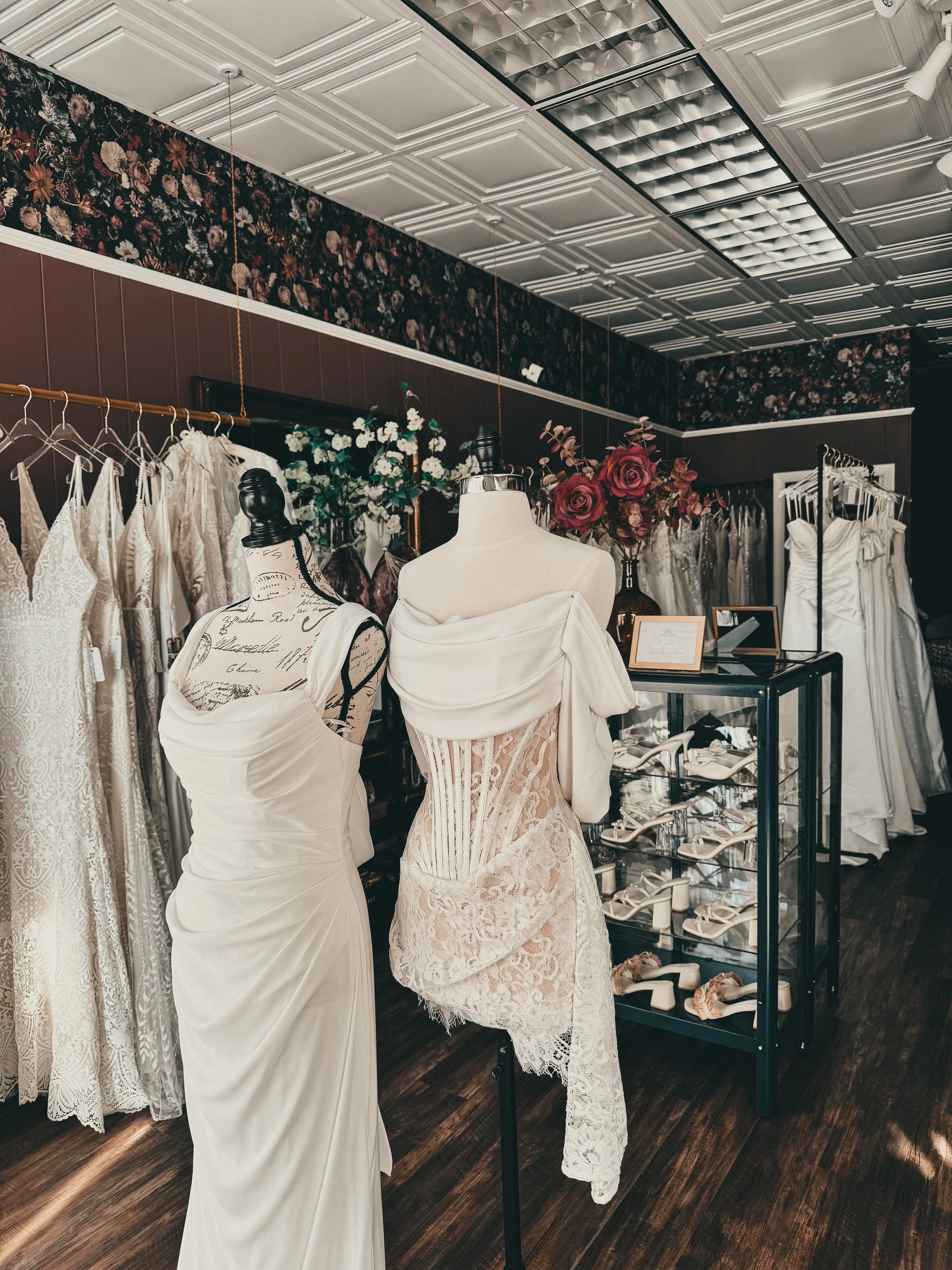 A bridal boutique displaying wedding dresses and shoes, with mannequins showcasing bridal gowns, floral arrangements in the background, and a glass shoe display case.