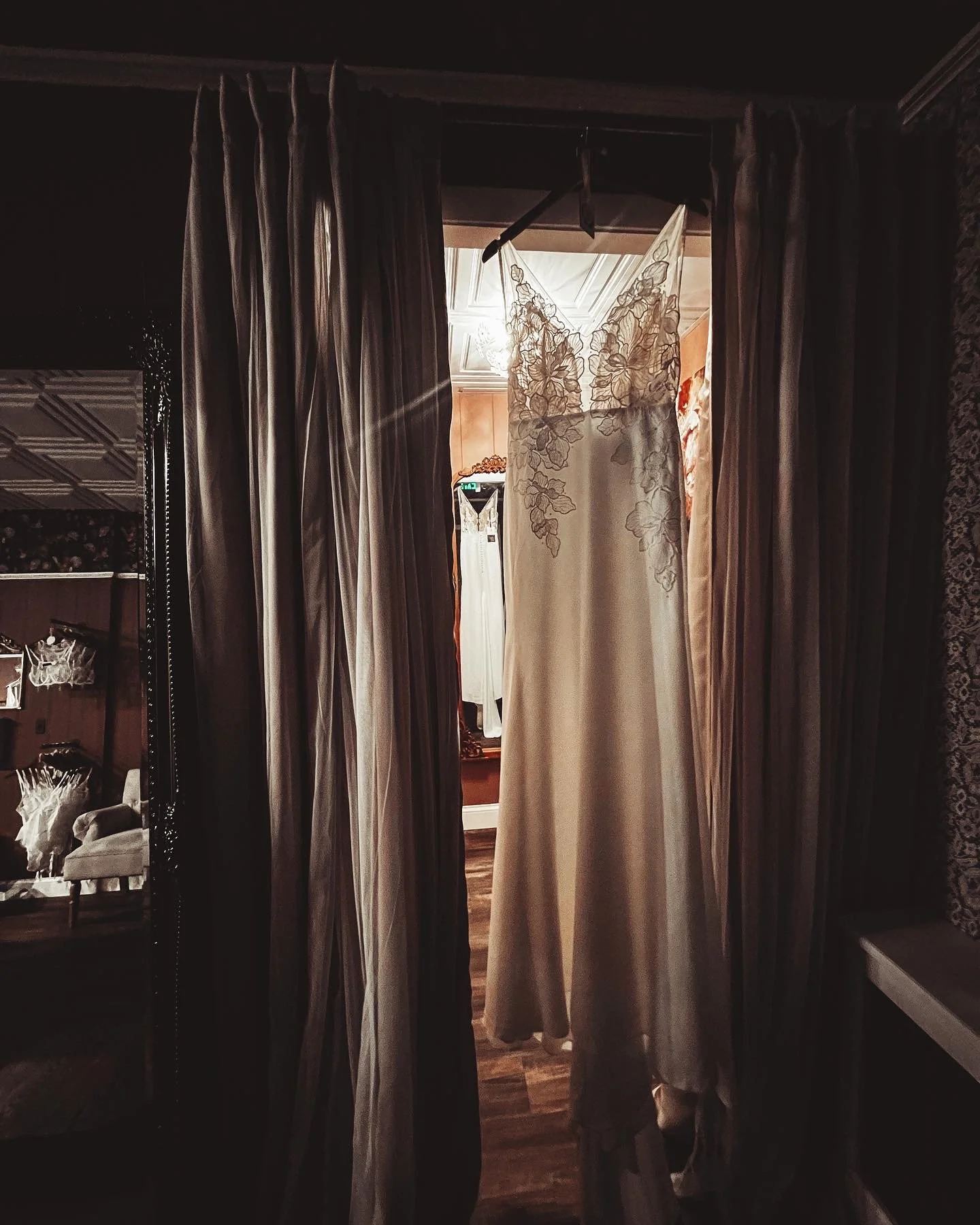 A wedding dress with lace detailing hanging behind curtains in a dressing room.