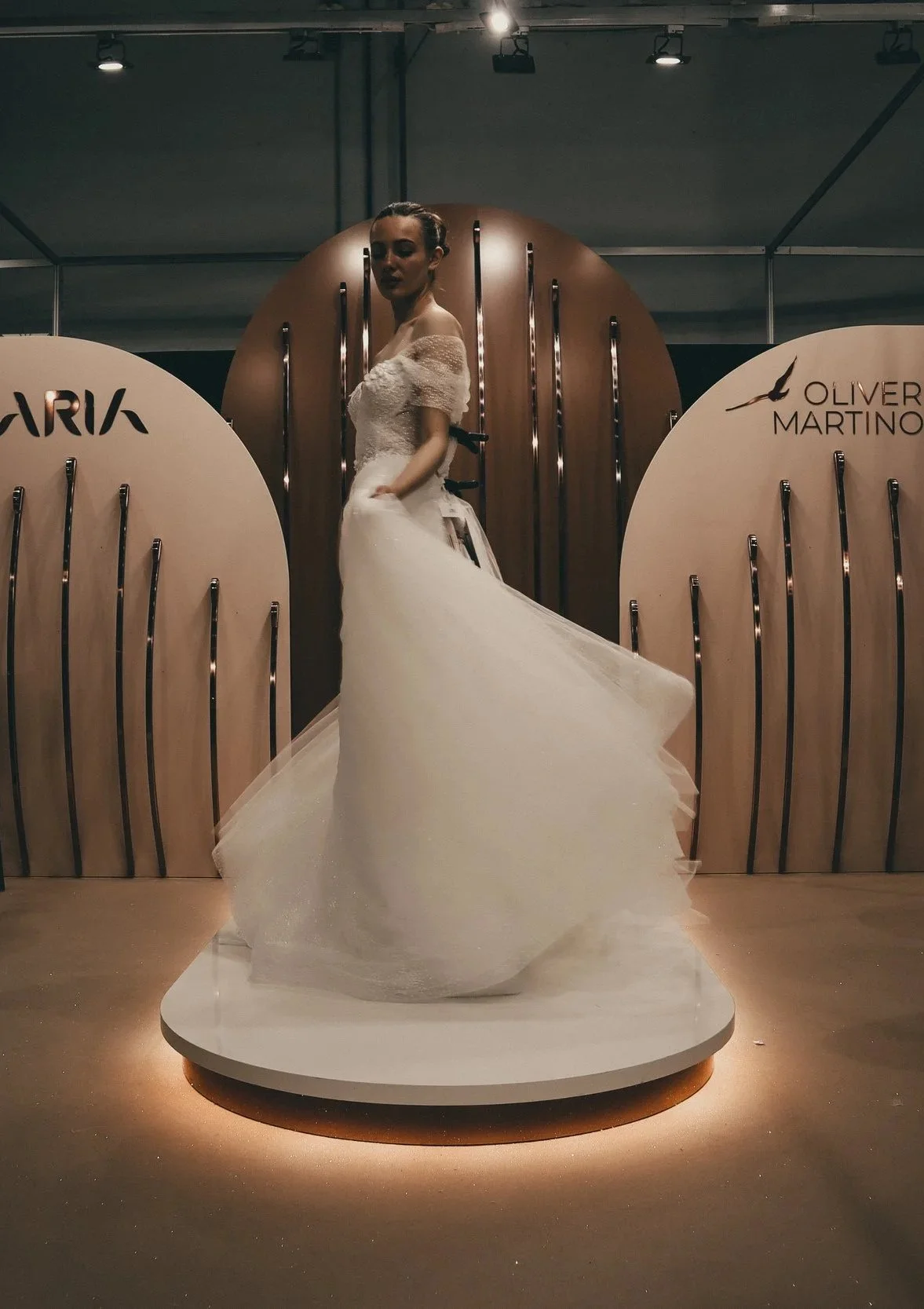 A woman in a wedding dress standing on a platform at a bridal exhibition, with a backdrop displaying the brands 'Mara' and 'Oliver Martino'.