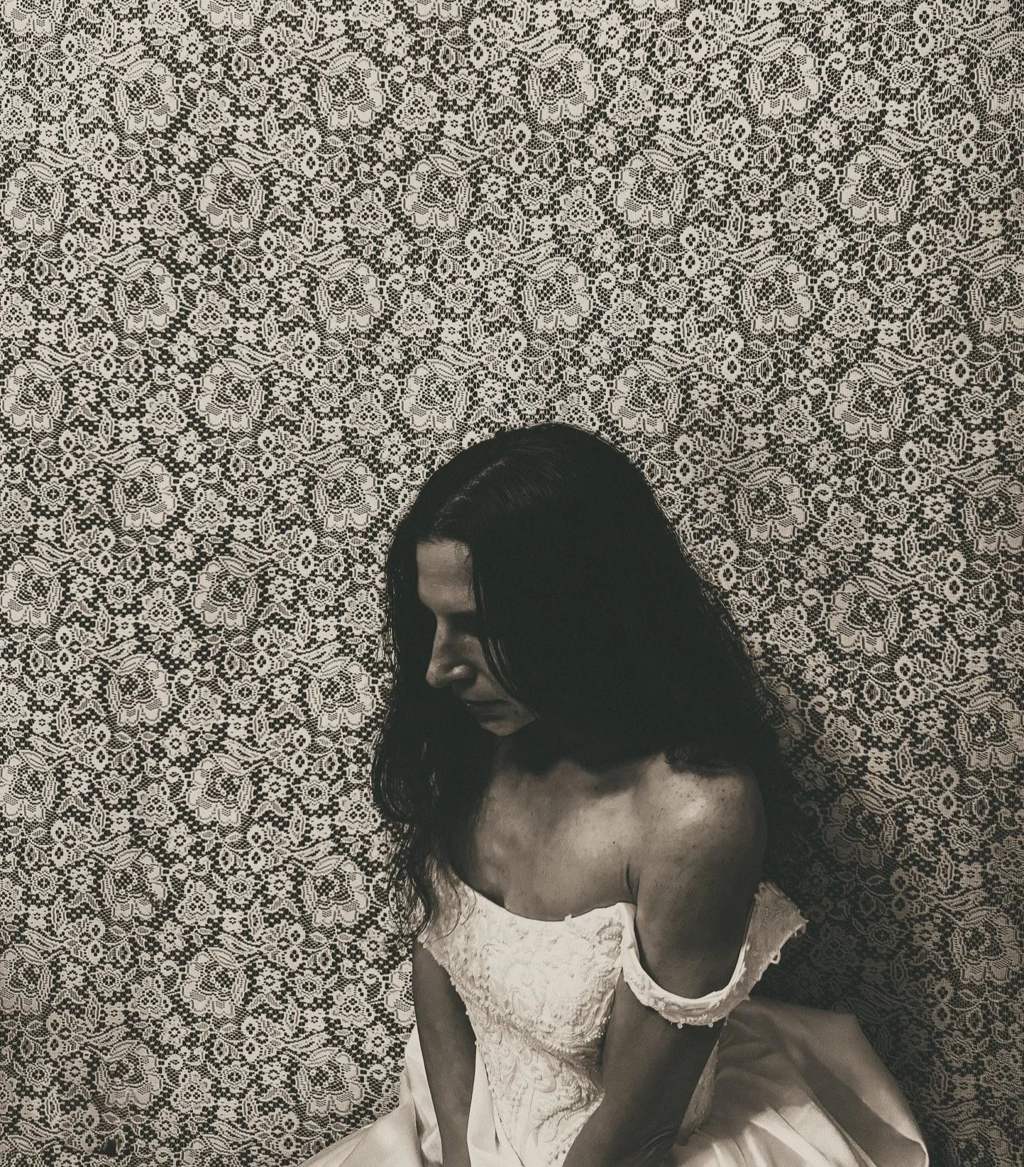 A woman with long dark hair wearing a white lace dress is sitting against a patterned lace wall, looking downward with her eyes closed.