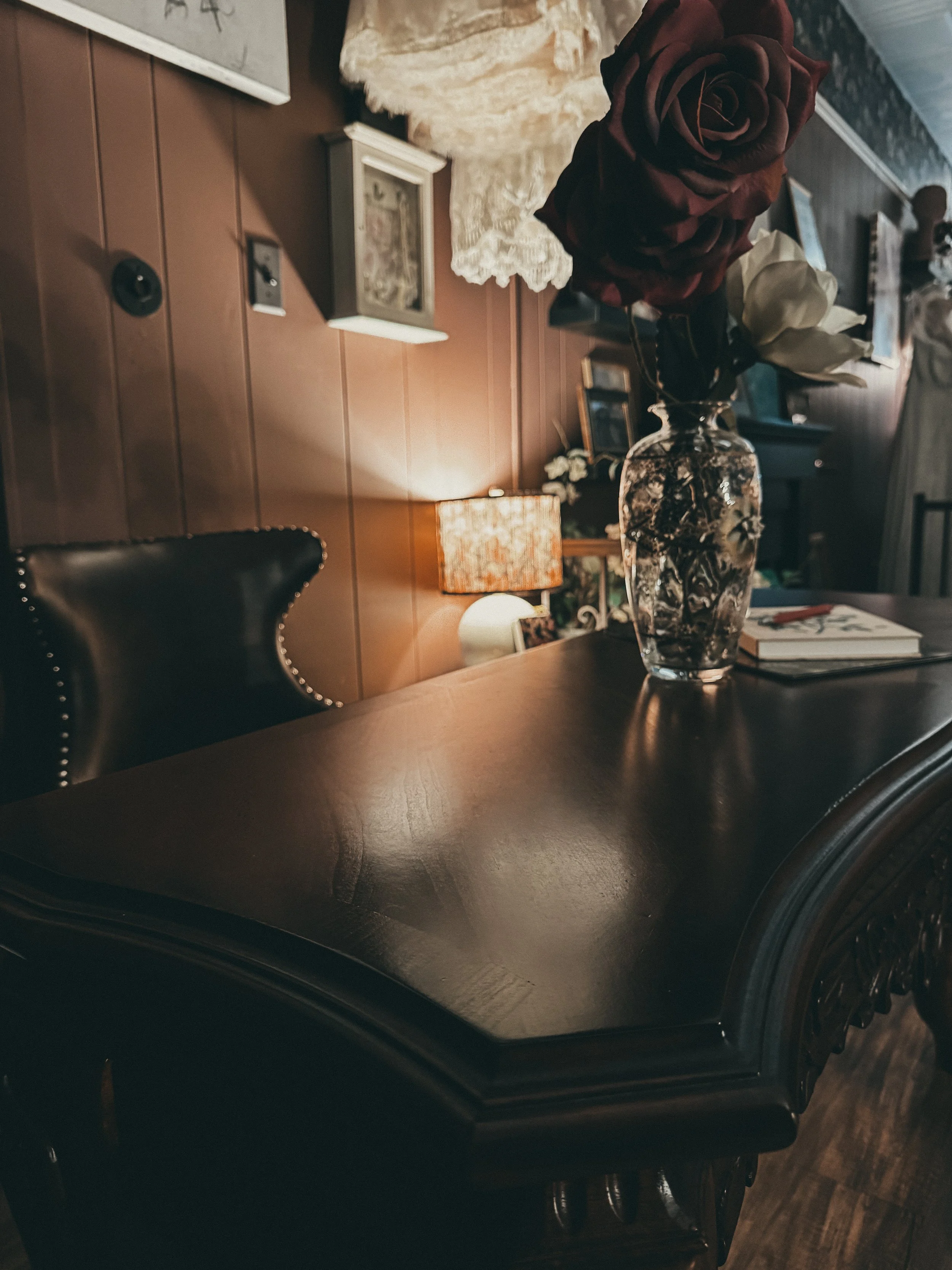 A dark wooden table with a glass vase holding red and cream artificial roses. In the background, there is a table lamp, framed pictures, and various decorative objects in a cozy room with wood-paneled walls.