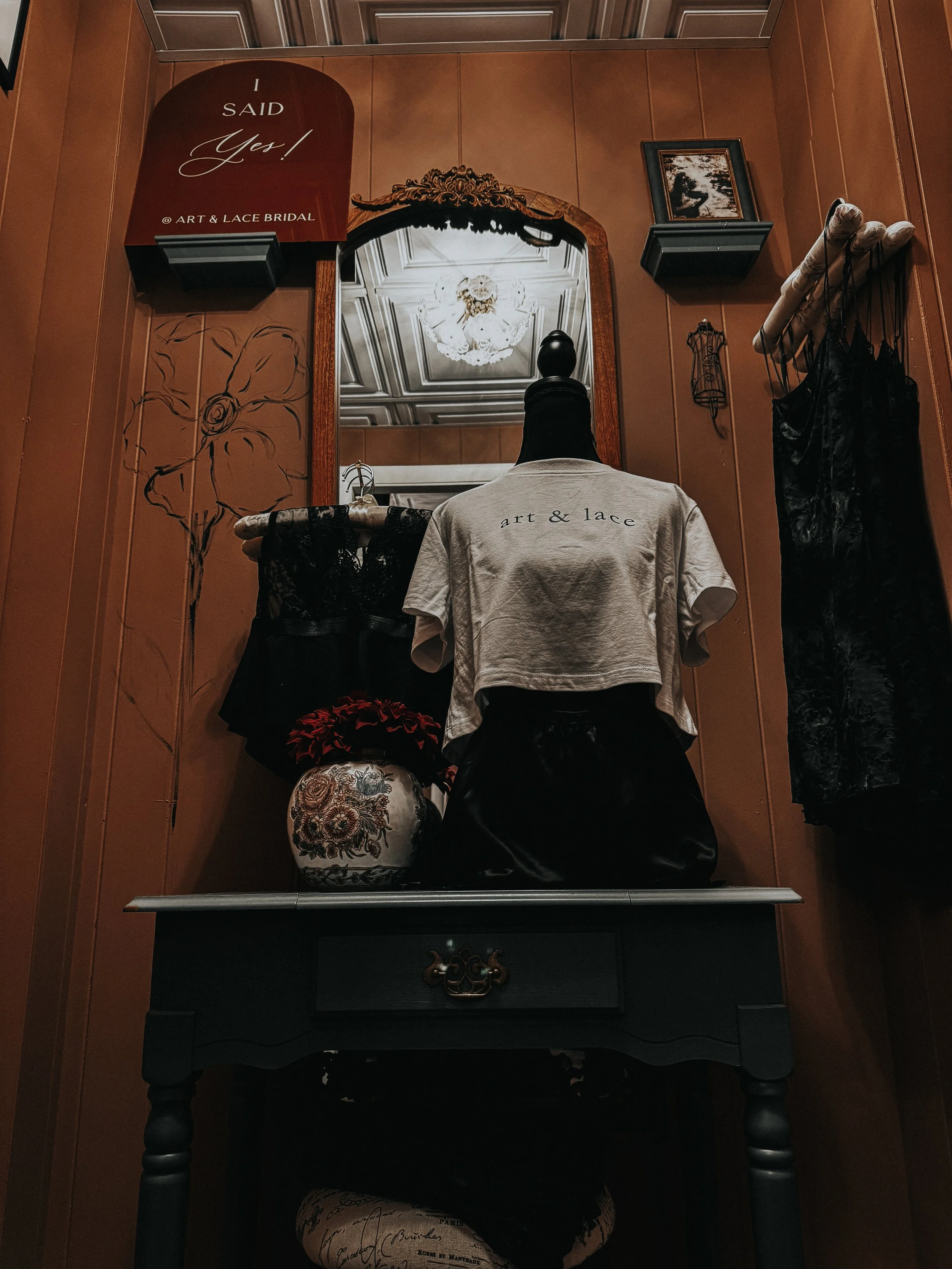 A black table with a decorative drawer handle against a brown wood-paneled wall. On the table, there is a mannequin torso wearing a knotted gray t-shirt with 'art & lace' written on it. Next to the mannequin, there is a decorative vase with floral em