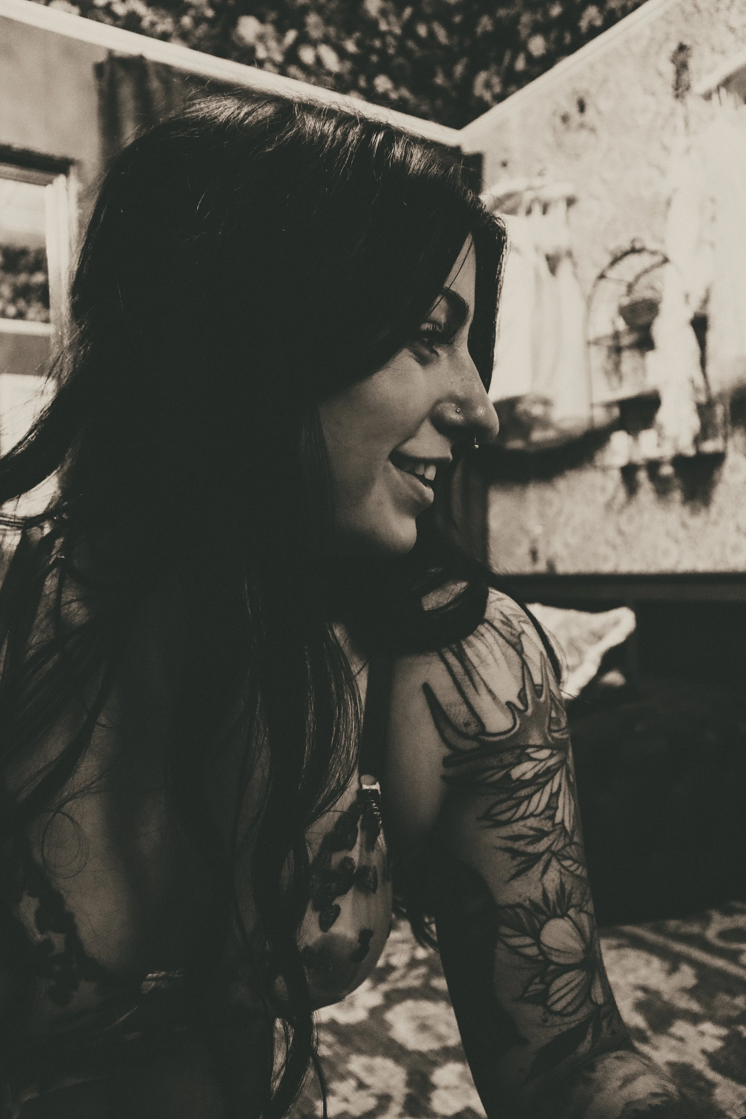 A woman with dark hair and tattoos on her arm, smiling while looking down, in a room with vintage decor and patterned rugs.