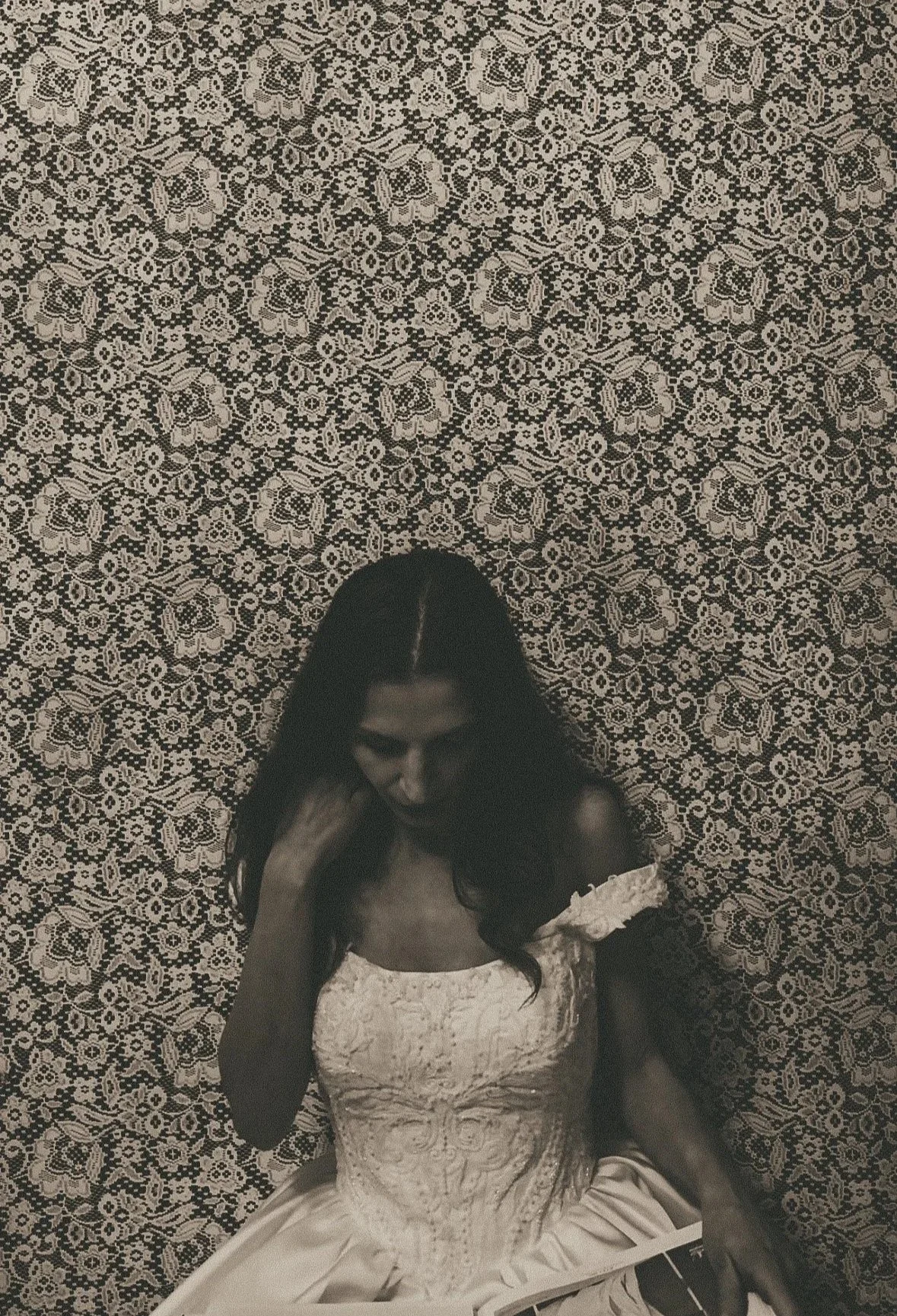 A woman in a light-colored, ornate dress sitting on a patterned floor, with her head bowed and hand touching her neck.