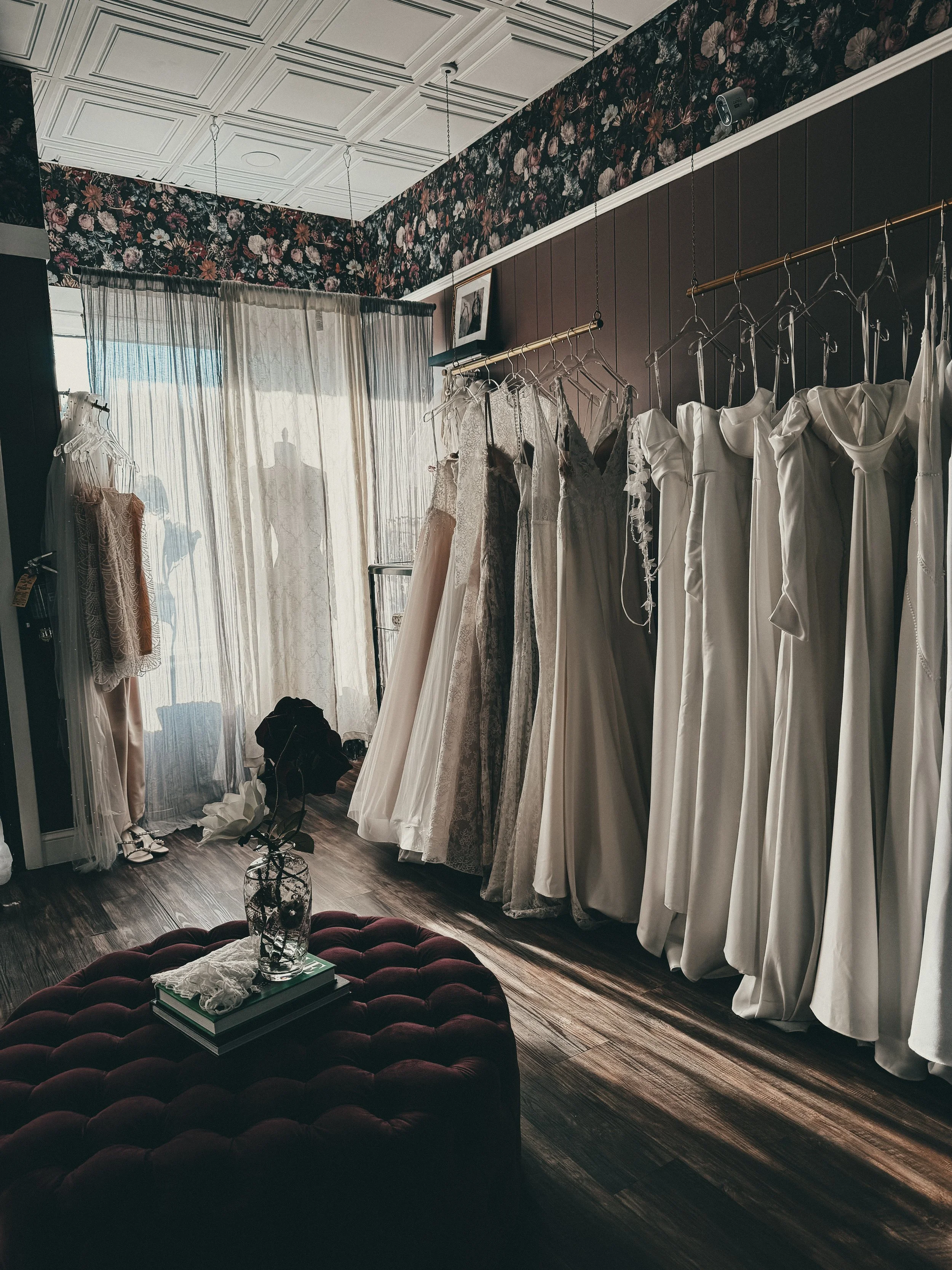 Bridal studio corner with floral wallpaper, burgundy tufted ottoman, and hanging romantic heirloom gowns at Art & Lace Bridal.