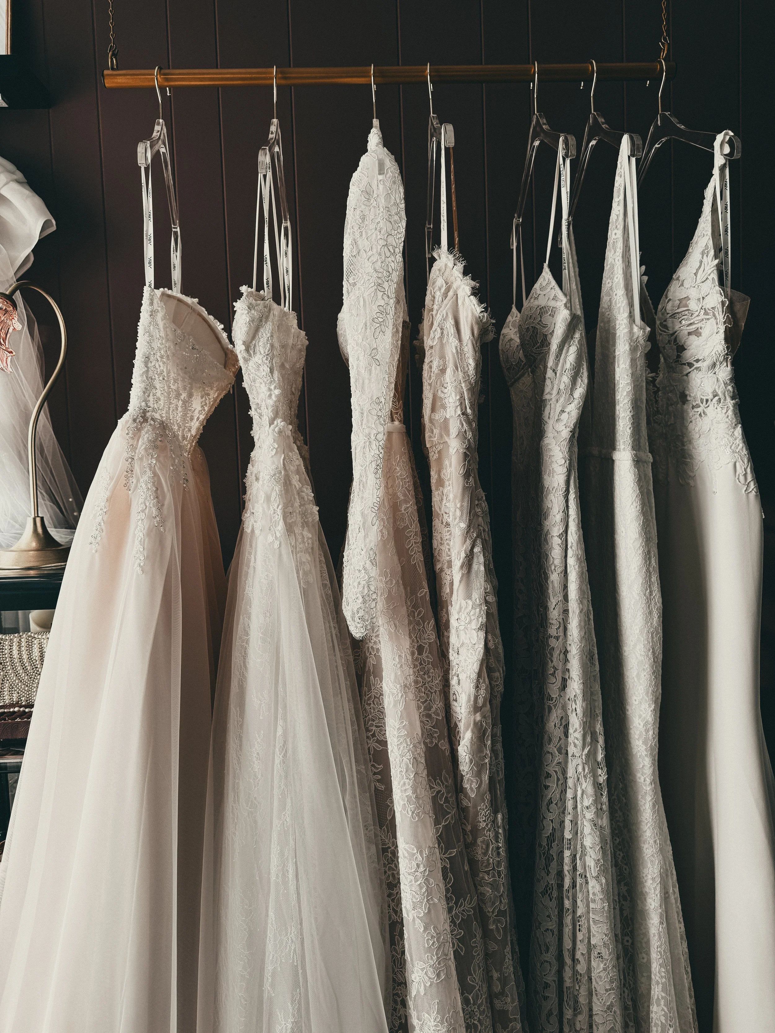 Curated rack of romantic heirloom wedding gowns under tin ceiling and floral wallpaper at Art & Lace Bridal, Somerset, MA.