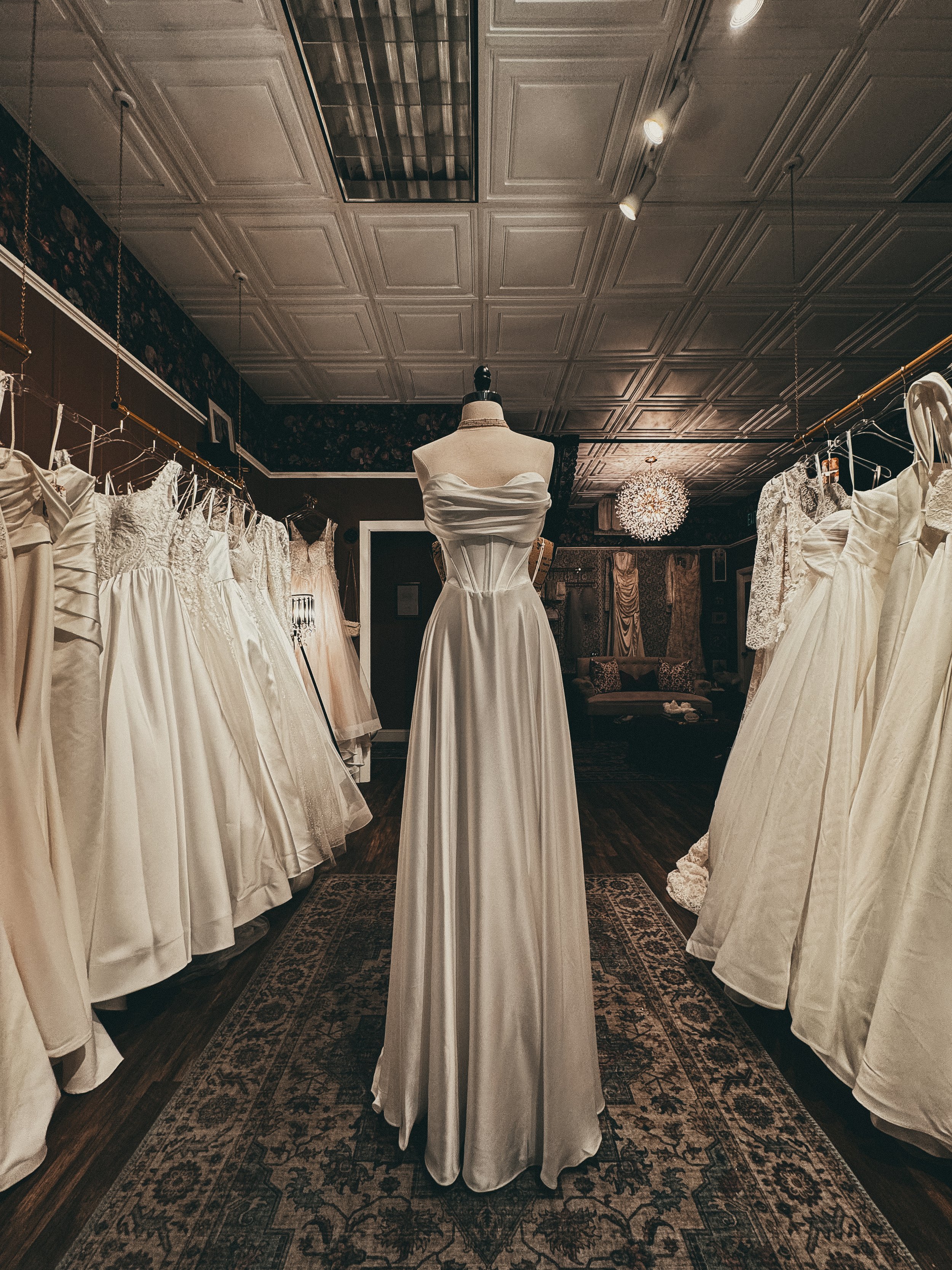 A wedding dress store with a mannequin wearing a strapless satin wedding gown, surrounded by other wedding dresses hanging on racks, with a vintage rug on the wooden floor and ornate ceiling tiles.