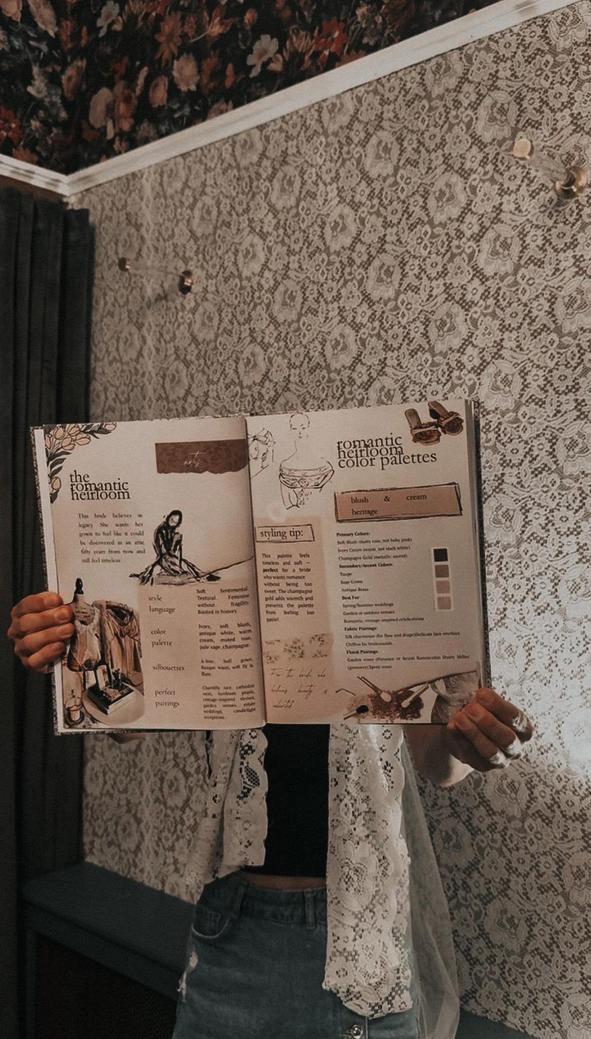 Person holding an open magazine featuring fashion and interior design, with a lace-patterned wall and floral wallpaper background.