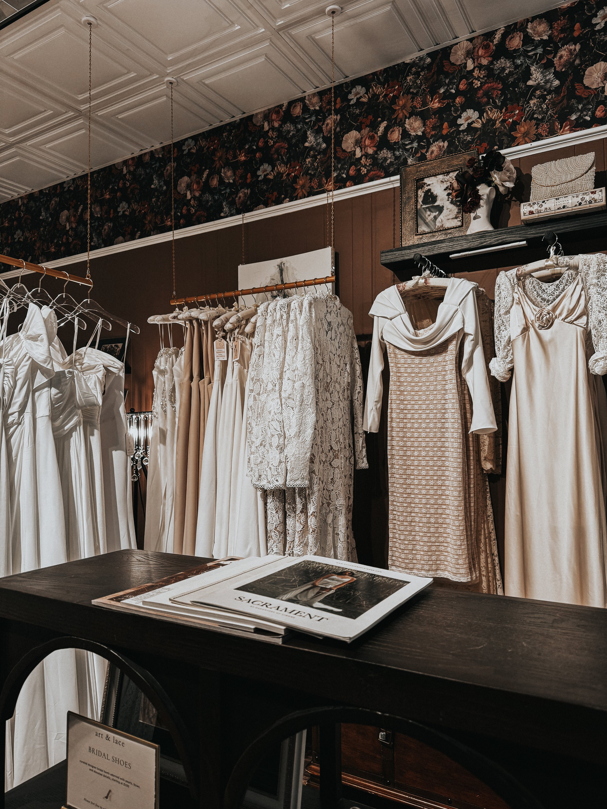 A display of vintage wedding dresses and formal gowns in a boutique, with a dark floral wallpaper accent and a wooden counter with magazines on it.