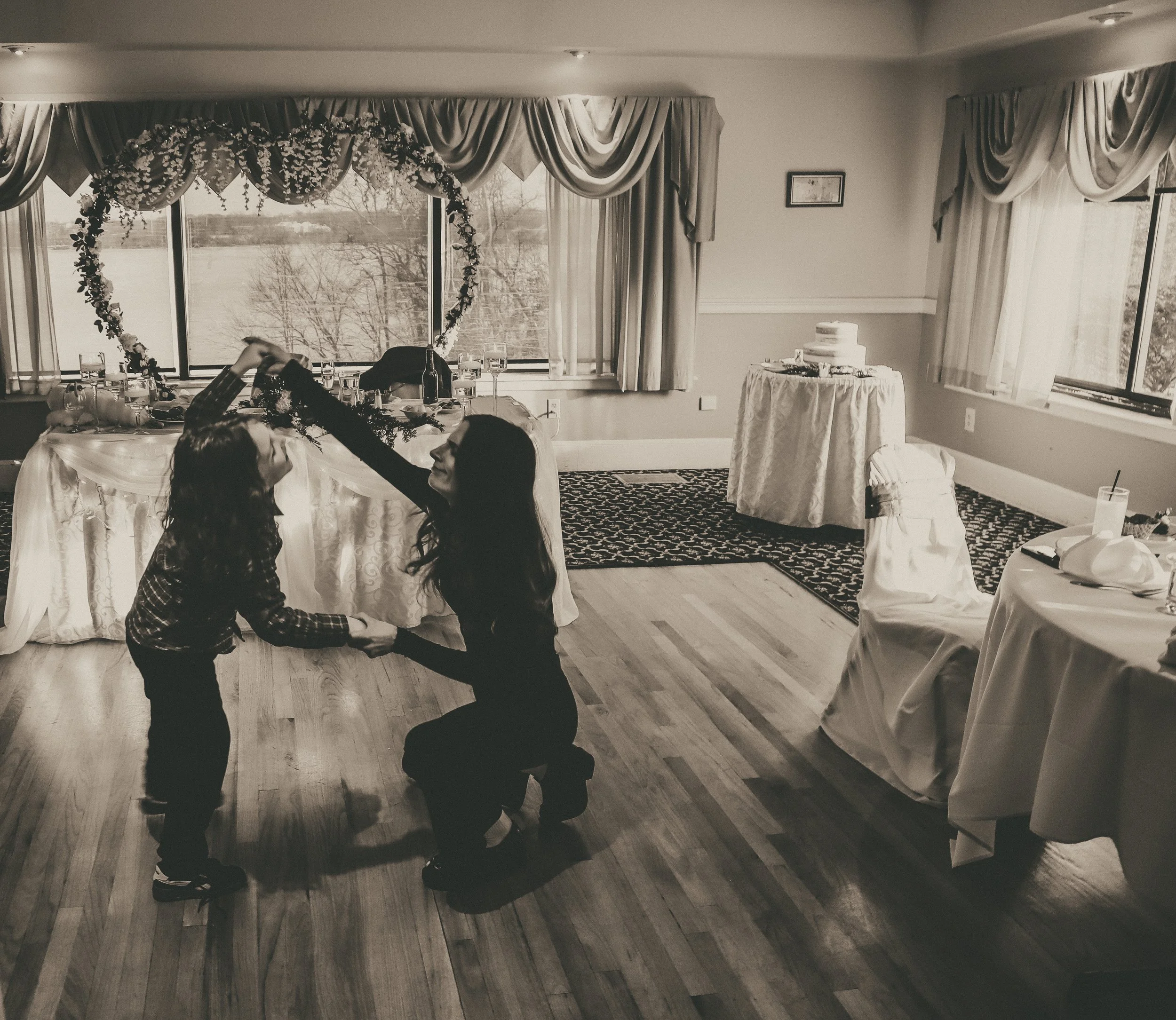 Two women dancing together at a celebration, with a decorated heart-shaped floral arch and a table with a wedding cake in the background.