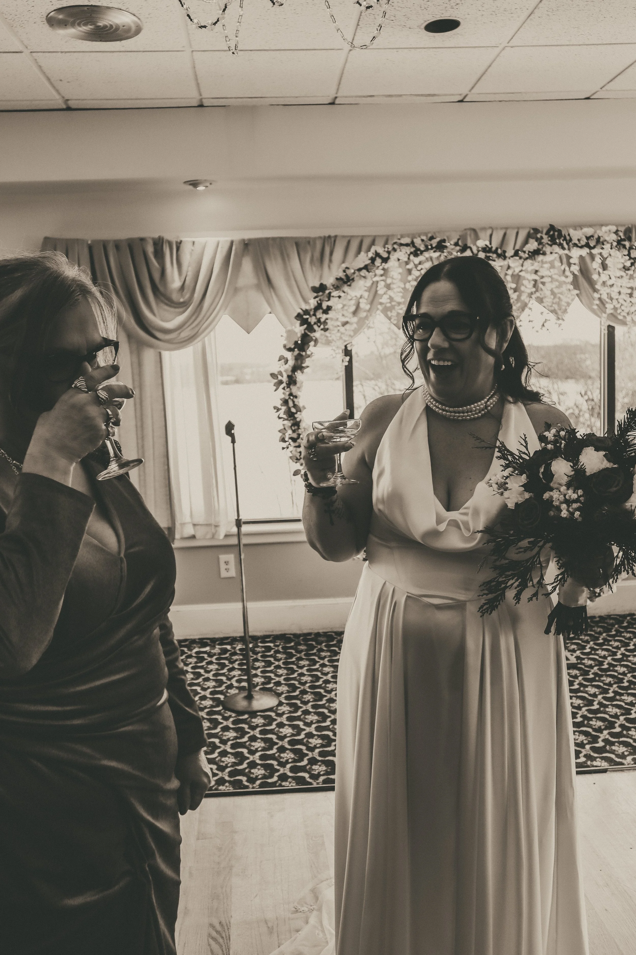Two women celebrate and toast at a wedding reception. The woman on the right, wearing a white dress, pearls, and glasses, holds a bouquet and a drink, smiling joyfully. The woman on the left, wearing a dark dress and glasses, also drinks and smiles. The background features windows with curtains and a floral arch.