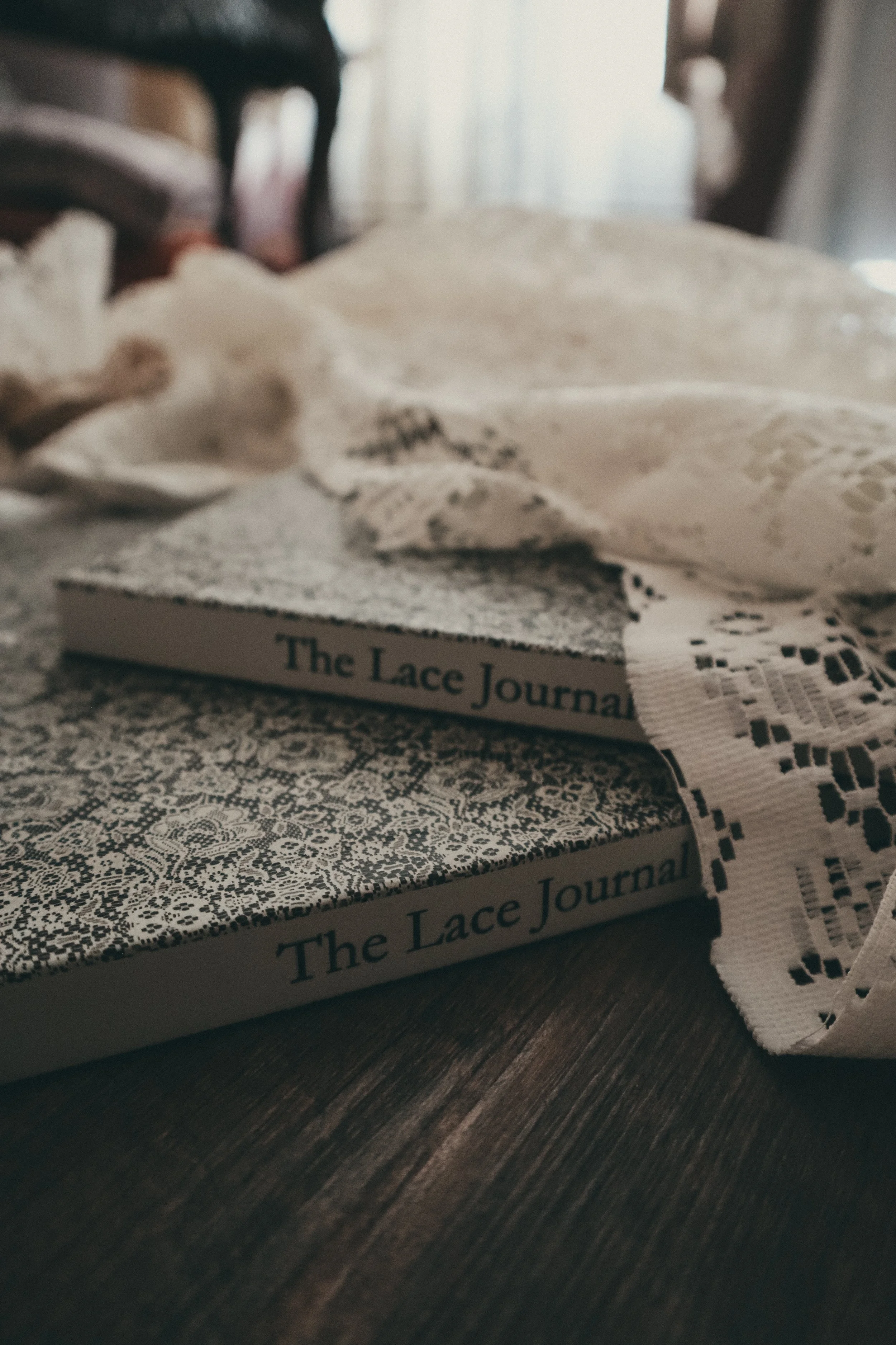 Two books titled "The Lace Journal" stacked on a wooden surface, with lace fabric and a lace ribbon partially visible on top of and beside them.