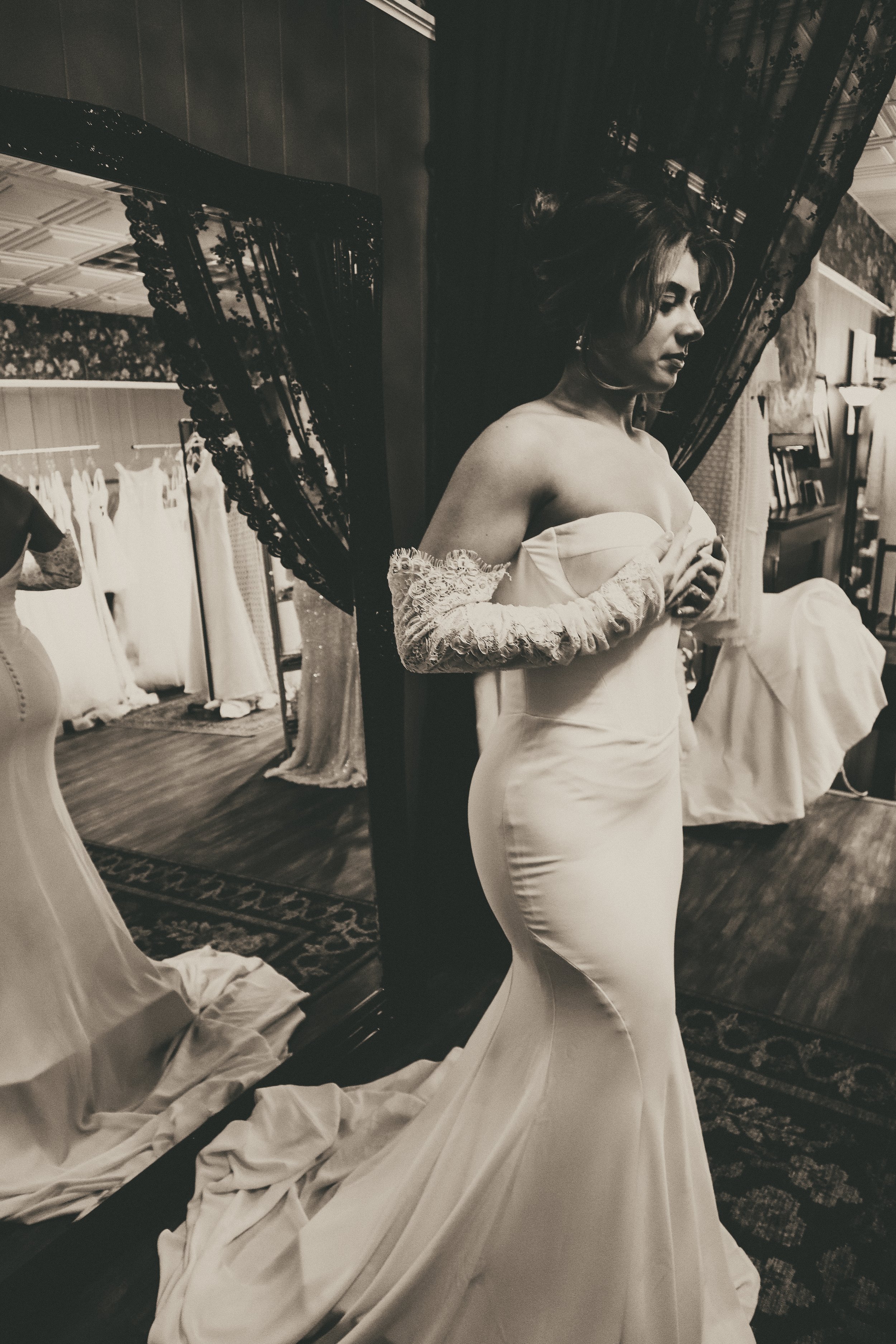 A woman in a wedding dress standing in a fitting room, looking at her reflection in a mirror. The dress is form-fitting with lace details on the sleeves and a long train on the floor.