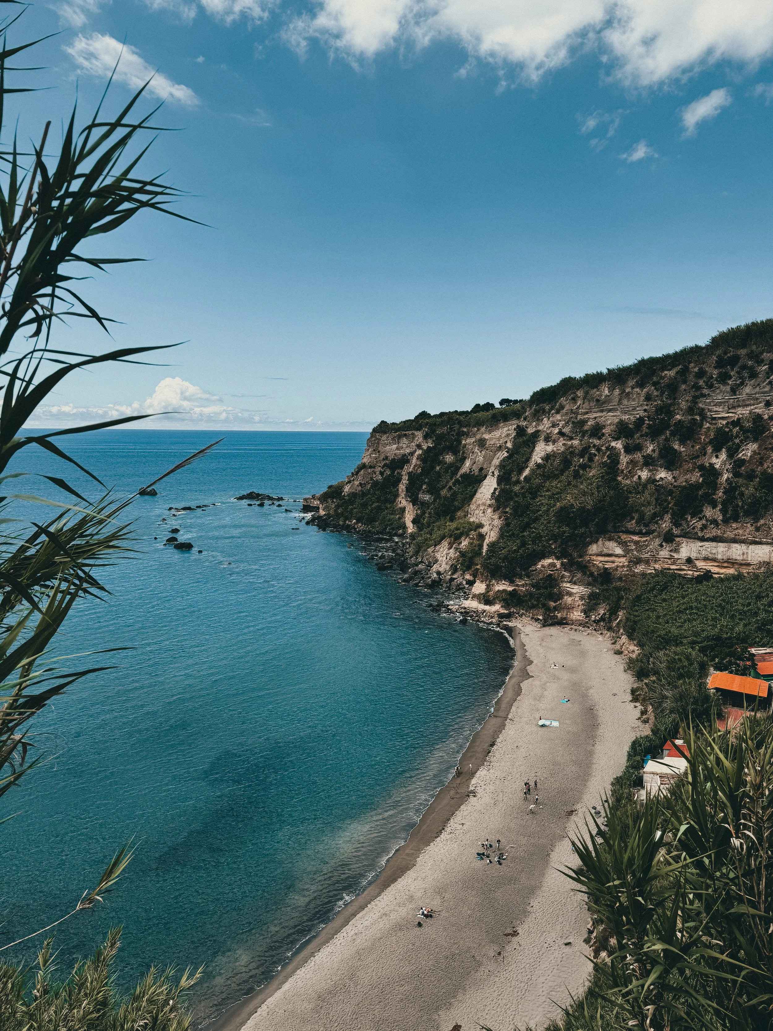 A panoramic view of a beach with a sandy shoreline, calm blue water, and tall cliffs covered with greenery. There are a few people relaxing on the beach, and some structures with orange and white roofs are visible near the edge of the image.