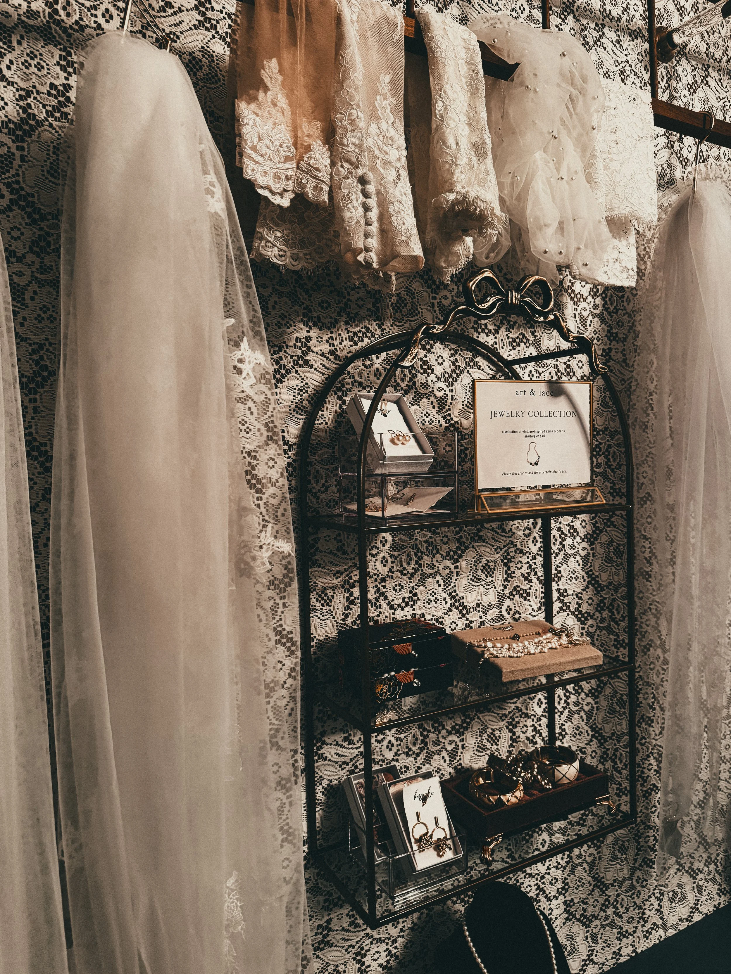 A display of vintage lace dress fabrics hanging on a rack, with a black metal jewelry stand below holding various jewelry pieces such as earrings, bracelets, and rings, against a lace-patterned backdrop.