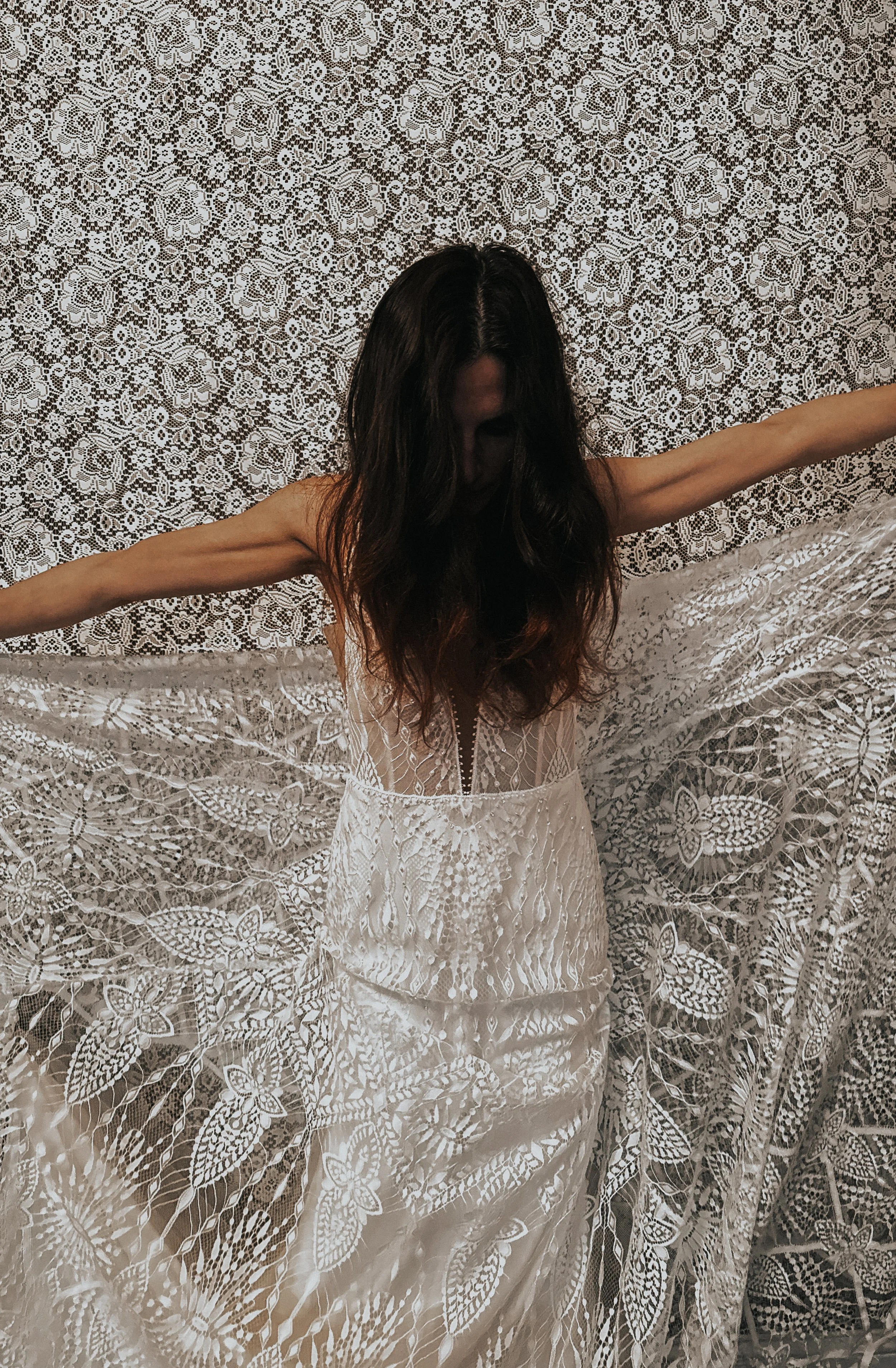 Woman in a white lace dress with arms outstretched, standing in front of a lace curtain background.