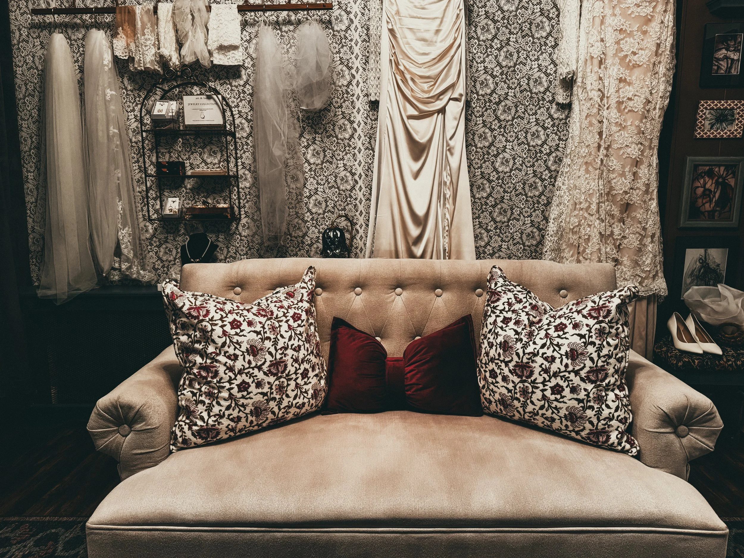 A vintage beige tufted sofa with three decorative pillows, two with floral patterns and one solid red, placed in front of a wall decorated with lace and satin fabric drapes, with wedding dresses hanging behind it and a small shelf displaying jewelry 