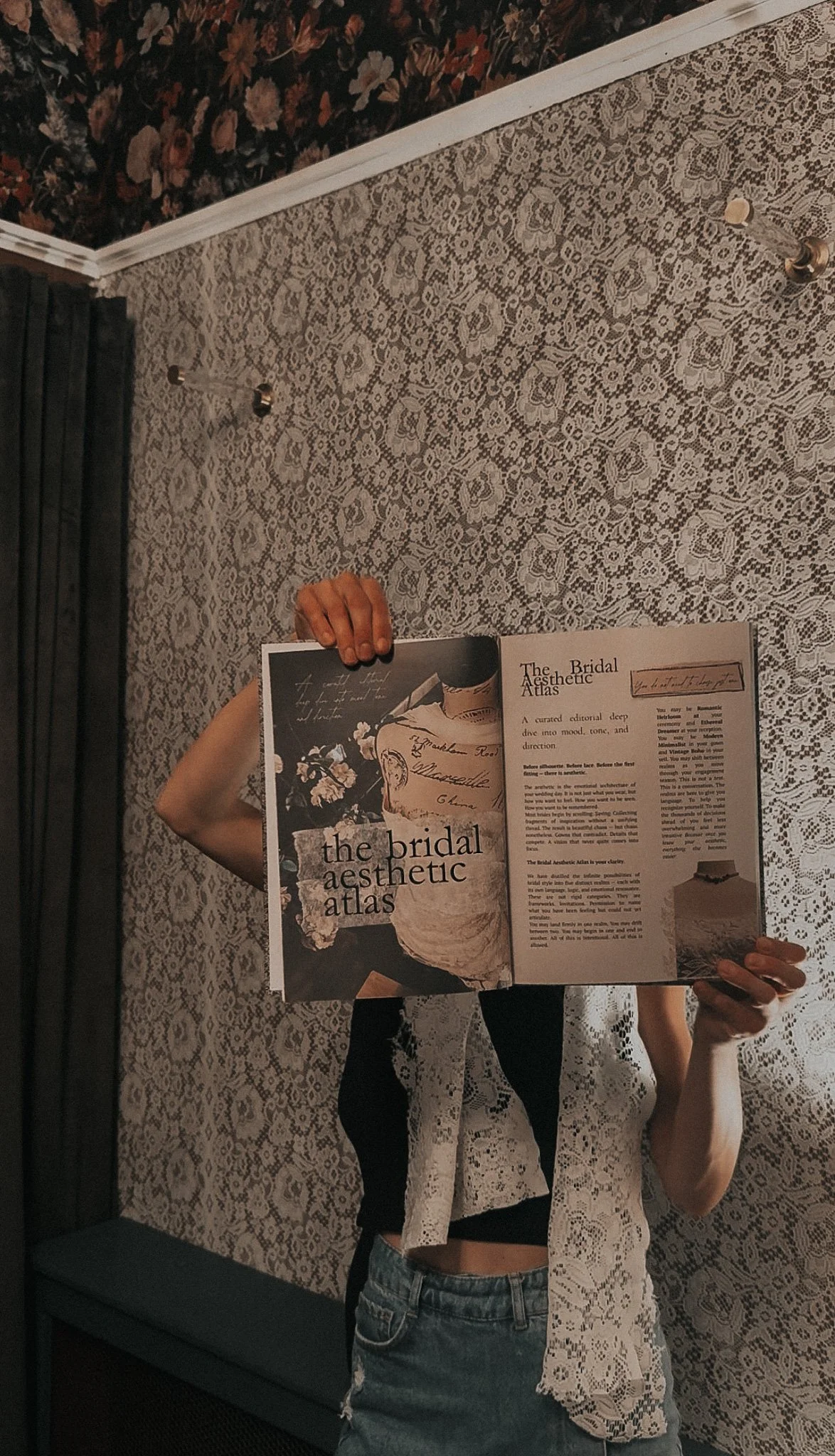 Person standing in front of a mirror, holding a magazine titled 'The Bridal Aesthetic Atlas' that covers their face. The person is wearing a white lace top and distressed jeans. The background features floral wallpaper and a lace curtain.