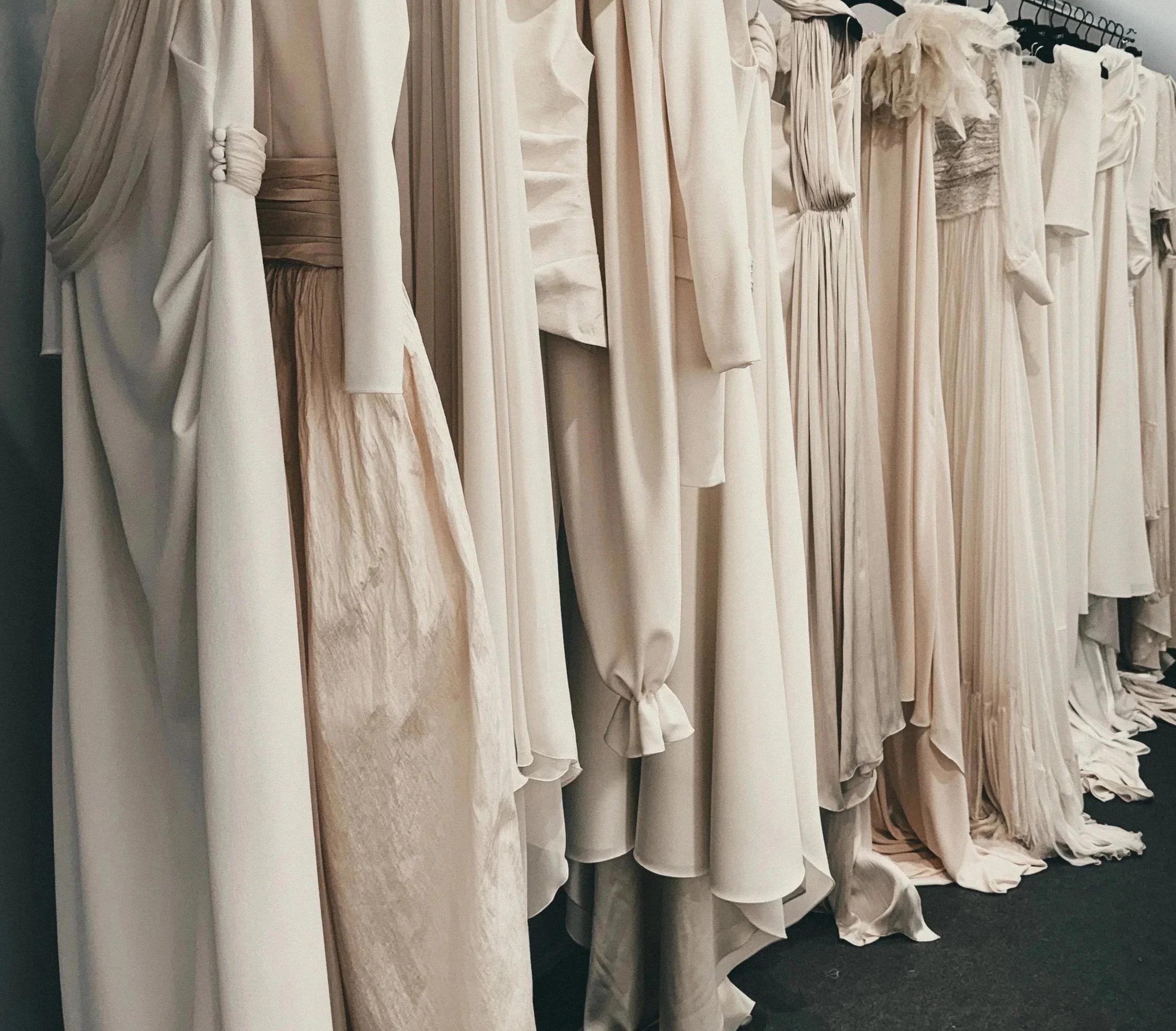 Clothing rack with various white and beige dresses hanging, some with pleats, ruffles, and tie details.